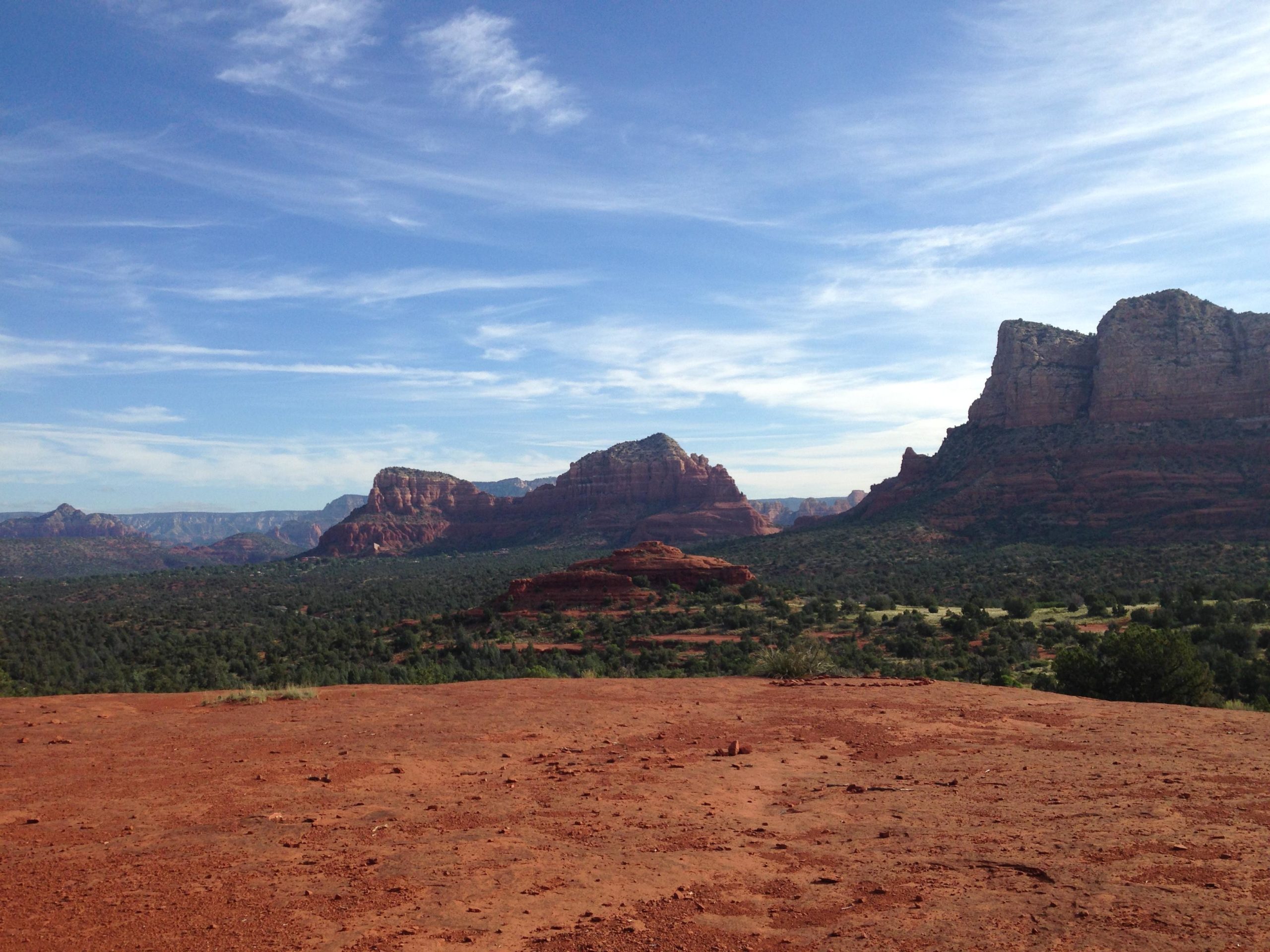 A scenic landscape featuring red rock formations under a clear blue sky, with green vegetation covering the valley below and distant mountains in the background. Baby Bell Trail mountain bike trail.