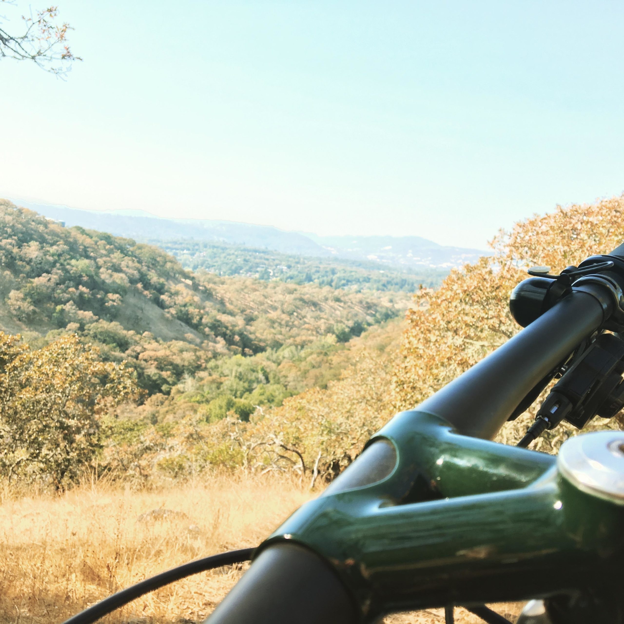 A close-up view of a bicycle's handlebars with a lush, green hillside and distant mountains in the background, under a clear blue sky. The landscape features trees and grasses, suggesting a scenic outdoor location ideal for biking or hiking. Annadel State Park mountain bike trail.