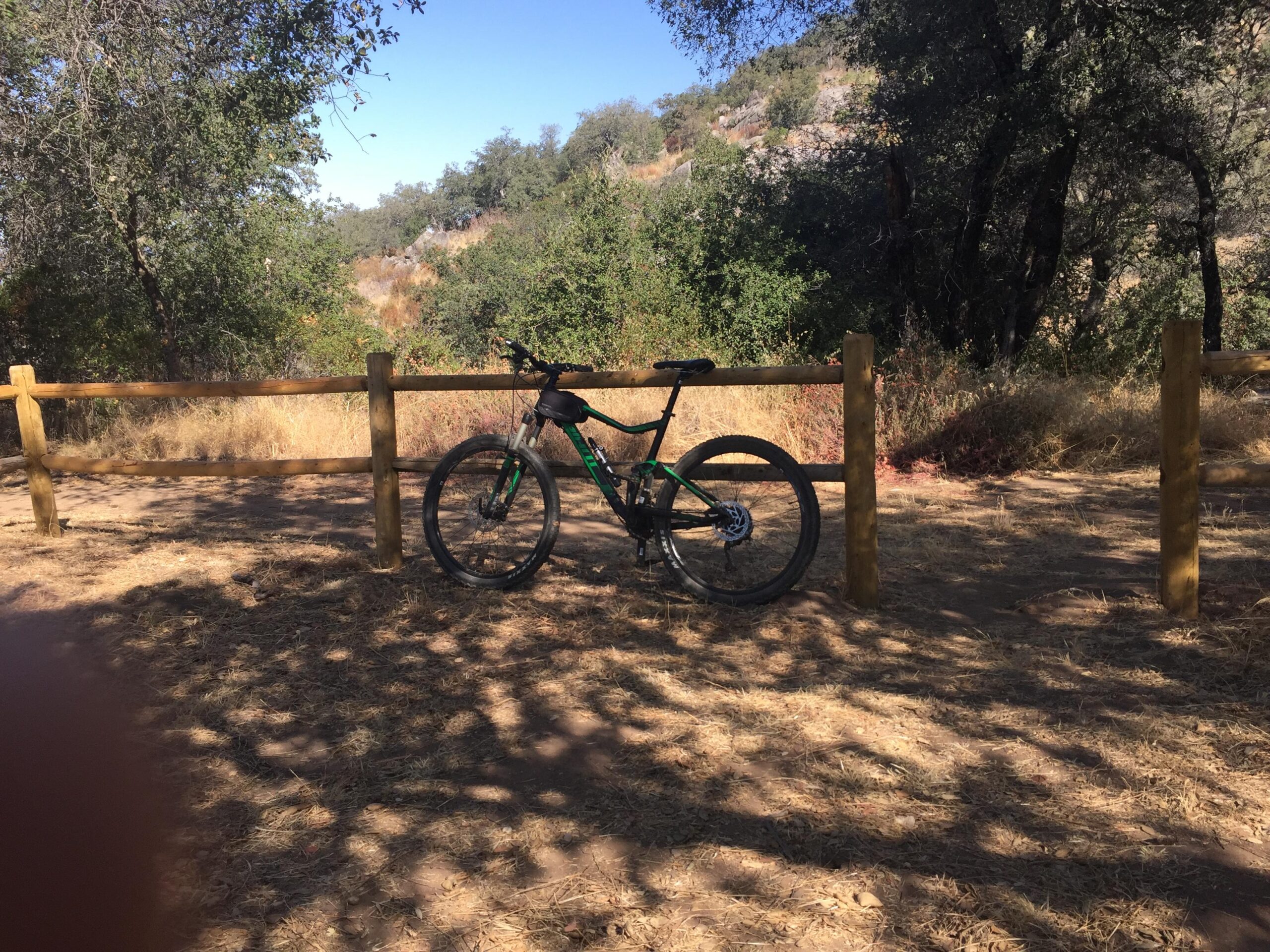 Giant Stance 27.5 2: A mountain bike leaning against a wooden fence, surrounded by trees and dry grass in a natural outdoor setting under clear blue skies.