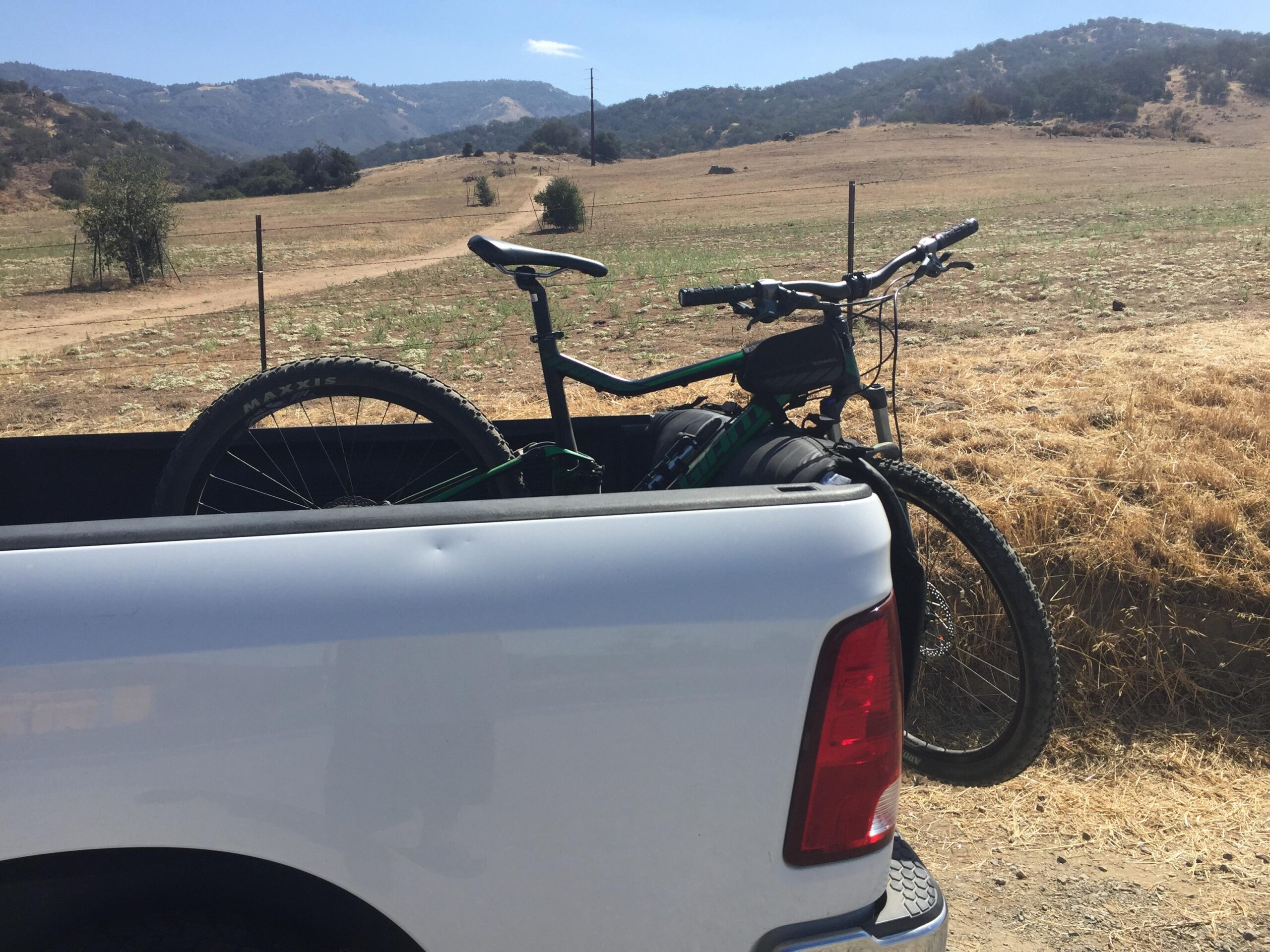 Giant Stance 27.5 2: A mountain bike resting in the back of a white pickup truck, parked near an open field with rolling hills in the background under a clear blue sky.