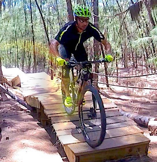 A mountain biker wearing a black and yellow outfit rides over a wooden bridge in a forested area. The biker is focused and in motion, surrounded by trees and natural scenery. Oleta River State Park mountain bike trail.