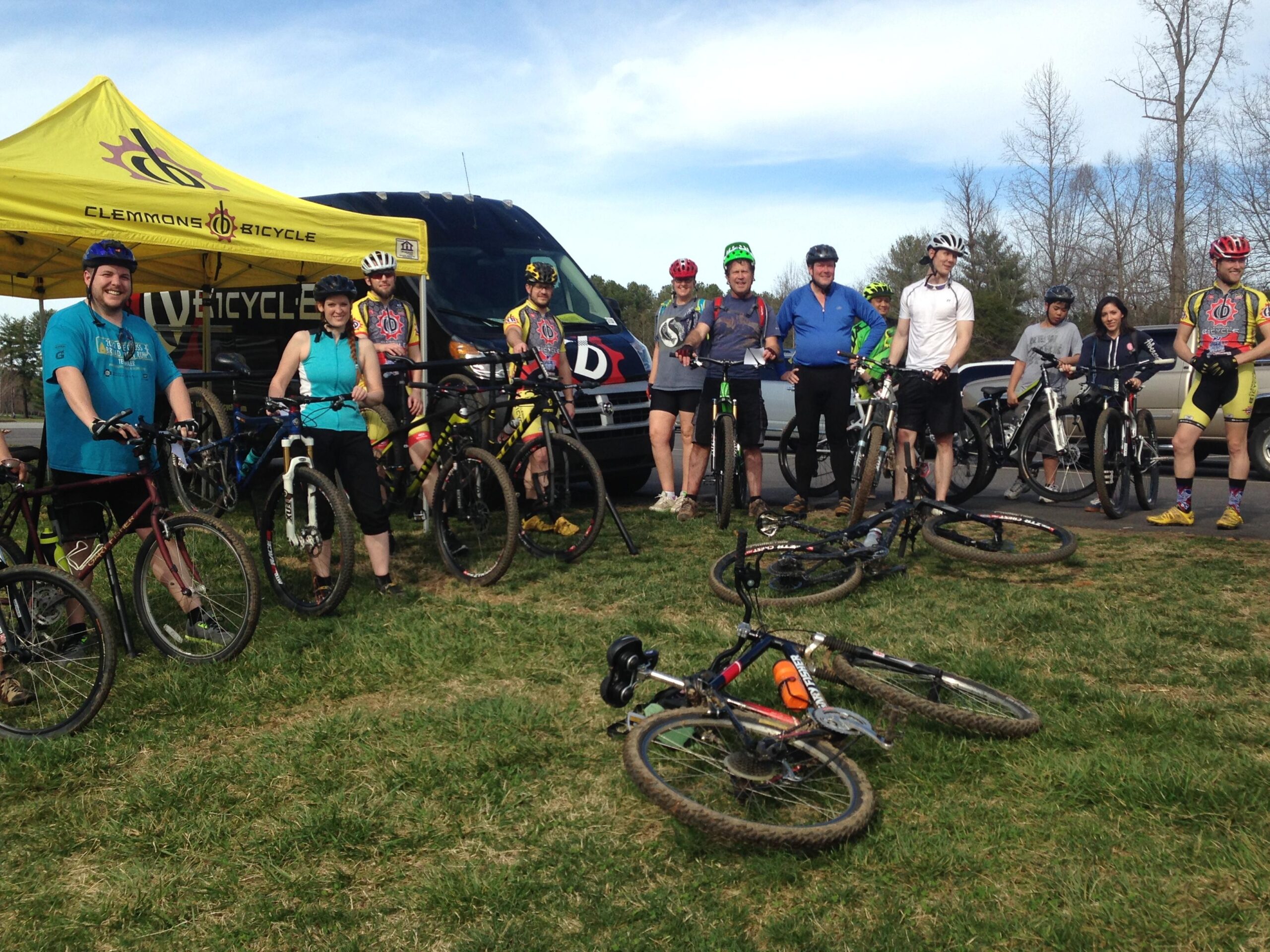A group of mountain bikers poses for a photo in front of a Clemmons Bicycle tent and van. The scene shows a mix of male and female cyclists, some on their bikes and others standing together, all wearing helmets and cycling gear. A few bikes are laid on the grass nearby, and the background features trees and a clear blue sky.