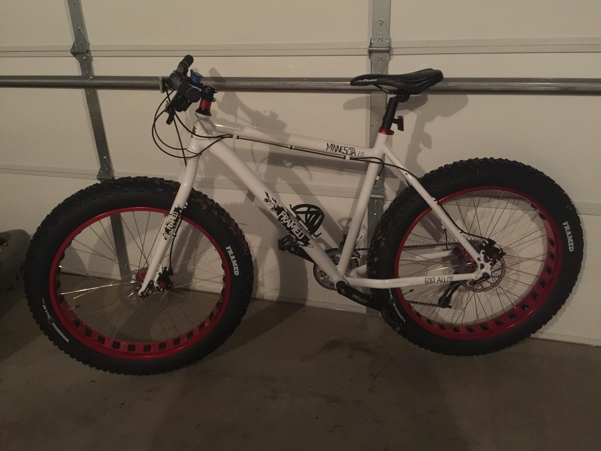 Framed Minnesota 2.0: A white fat bike with large, knobby tires featuring red rims, leaning against a garage wall. The bike's frame is labeled "Minnesota 2.0" and has a black saddle and handlebars. The garage has a concrete floor and metal support beams in the background.