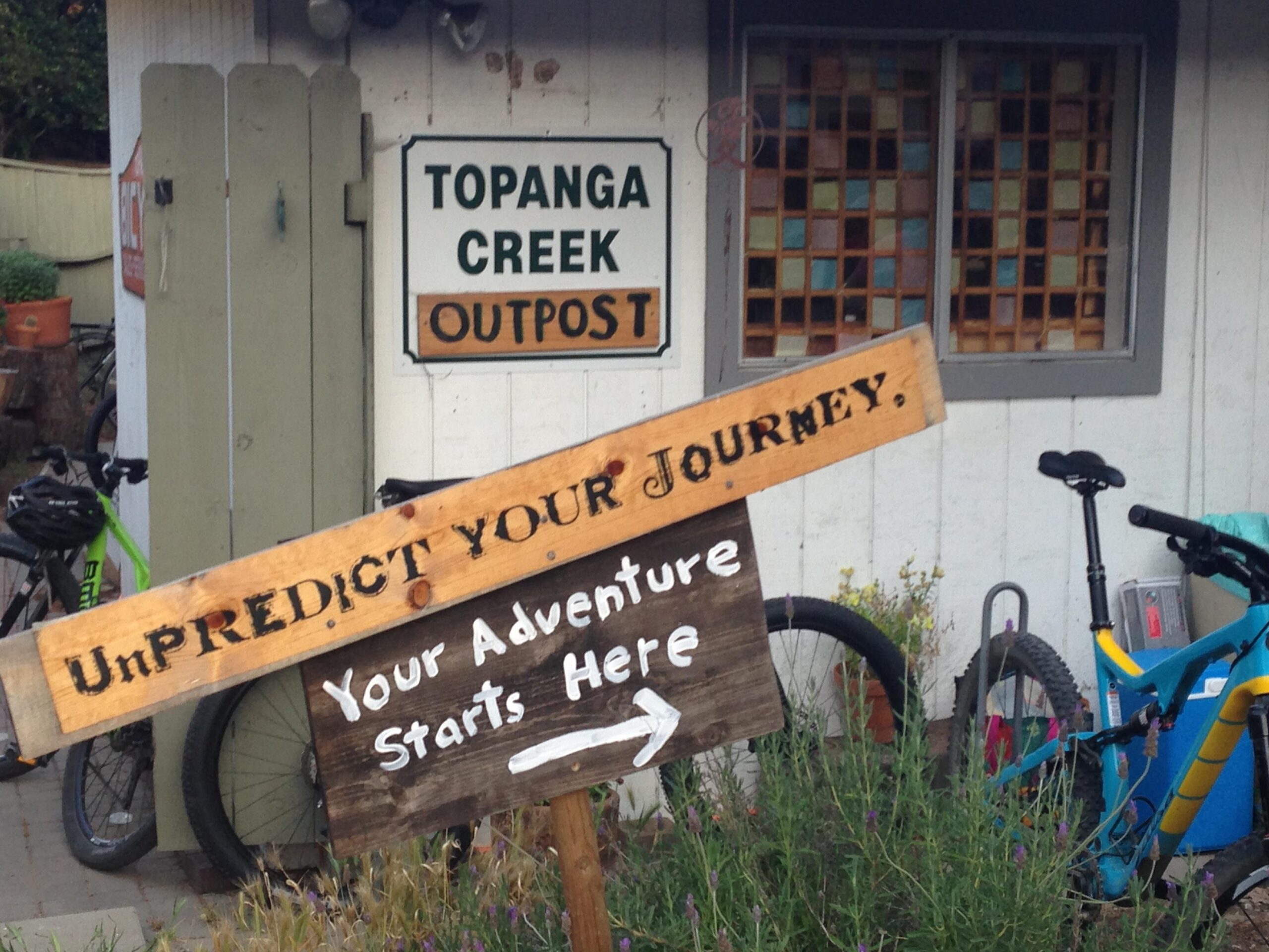 A rustic wooden sign reading "UnPREDICT YOUR JOURNEY" is prominently displayed in front of a building with a sign that says "Topanga Creek Outpost." The sign points to the right and states, "Your Adventure Starts Here." In the background, there are two bicycles parked beside the building, surrounded by greenery and flowers.