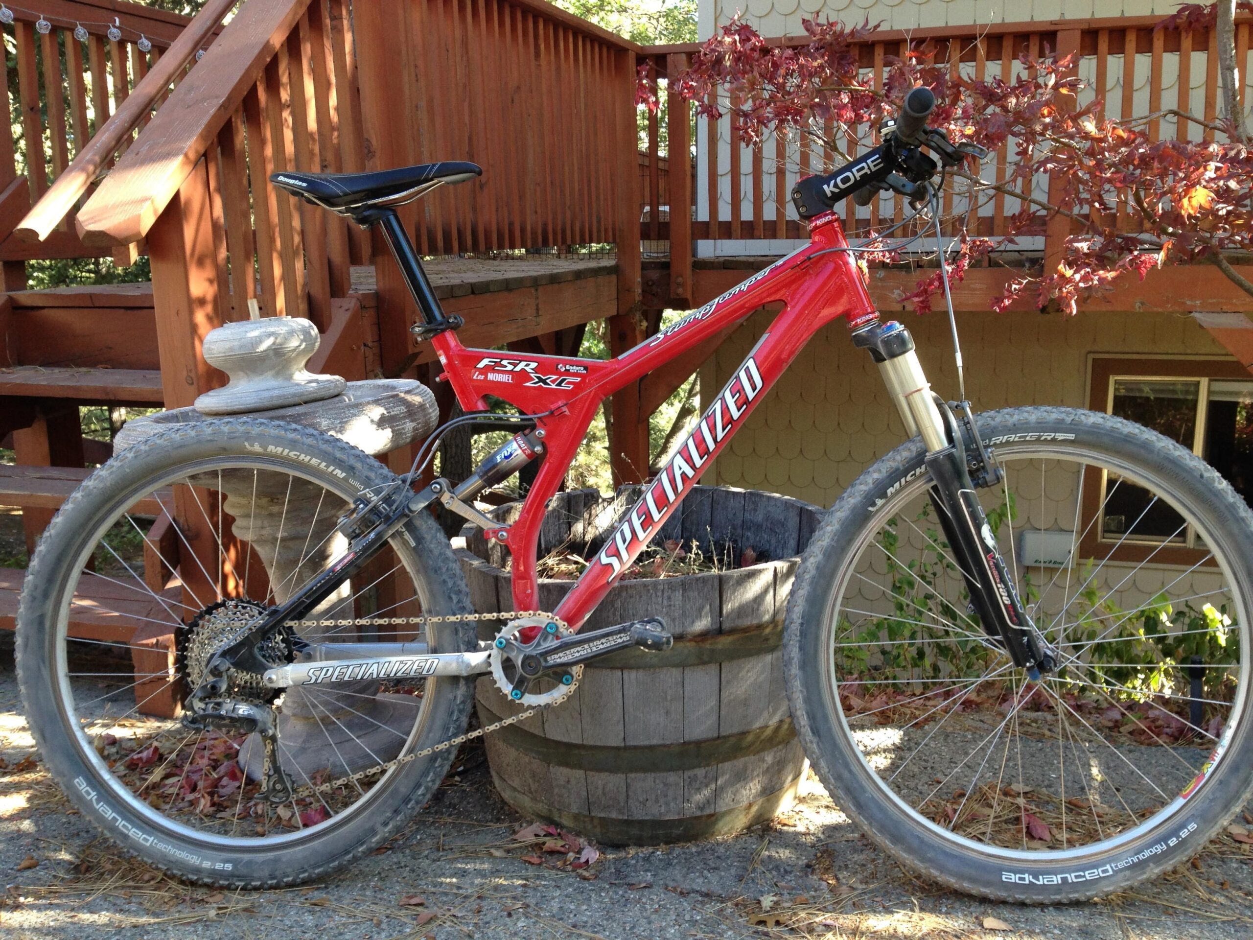 Specialized Stumpjumper FSR: A red mountain bike leaning against a wooden barrel, with a stone on top, in a garden setting. In the background, there is a wooden staircase and a house, partially obscured by a tree with red leaves. The bike features thick tires and a specialized frame, indicating it is designed for off-road use.