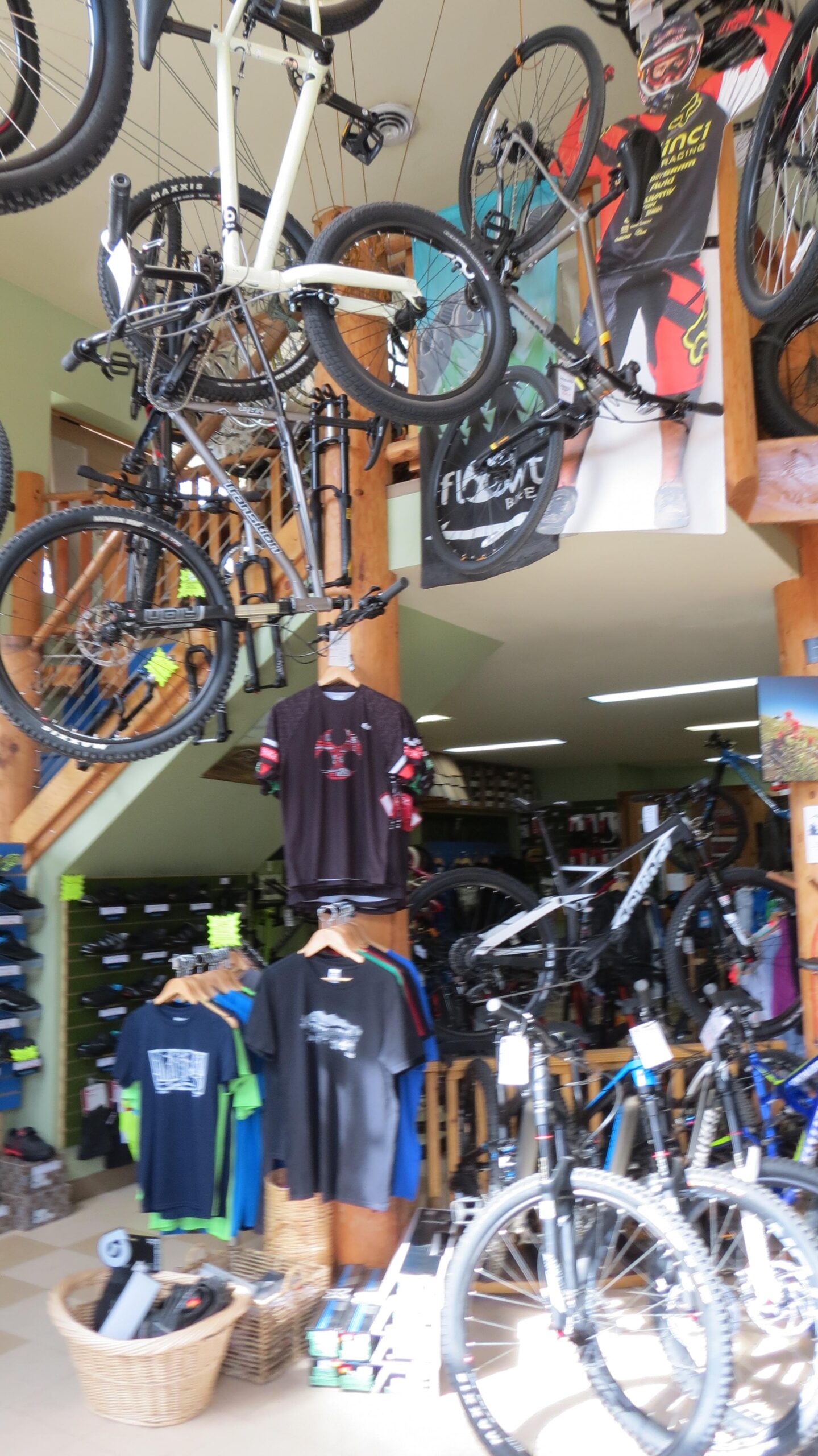 A brightly lit bicycle shop interior featuring various mountain bikes hanging from the ceiling and displayed throughout the space. The walls are adorned with colorful biking apparel, including jerseys and T-shirts, along with shelves stocked with biking accessories. A large poster of a cyclist is visible, and the shop has a warm wooden aesthetic.