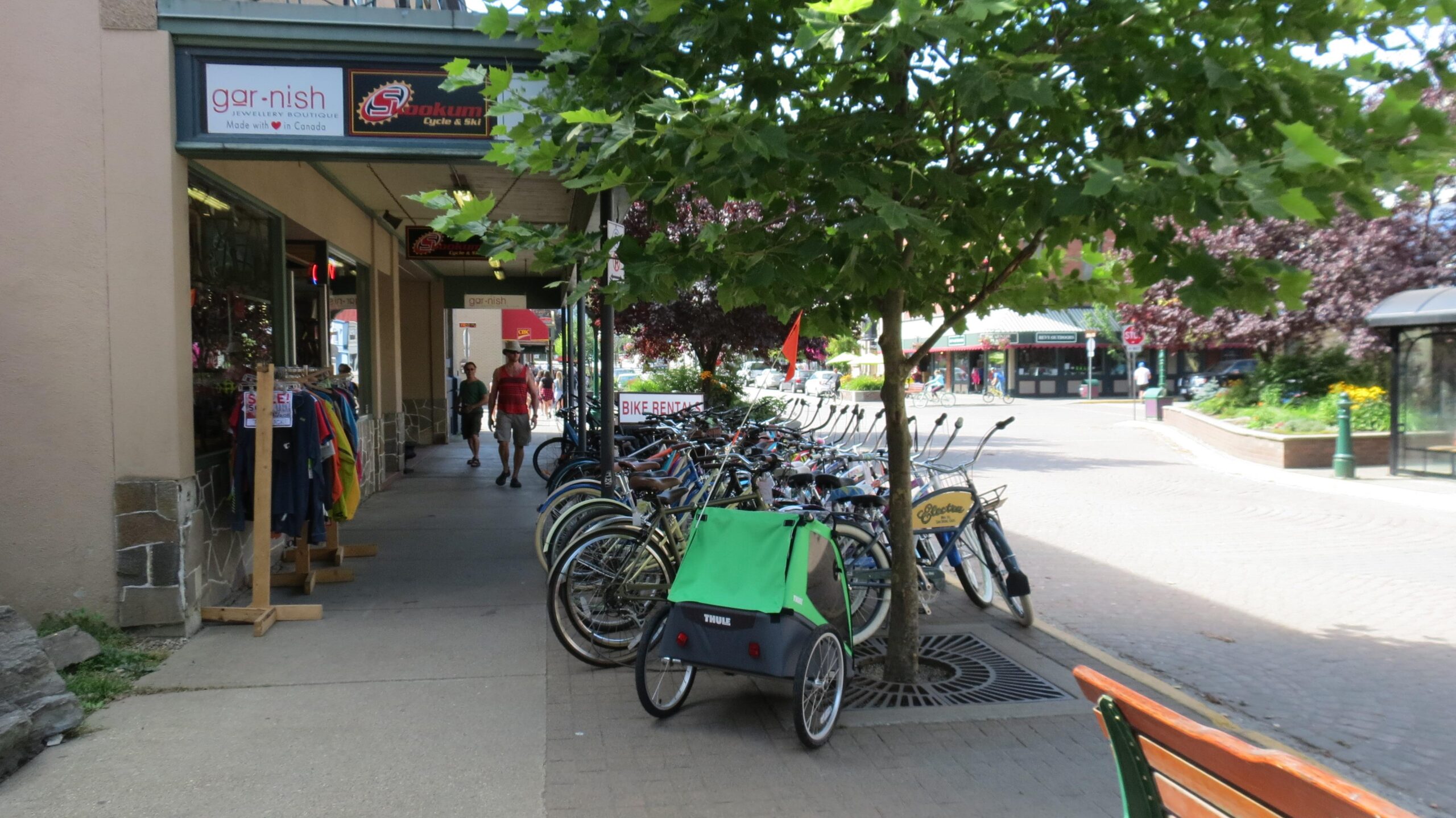 A bustling street scene featuring a row of parked bicycles alongside a sidewalk. A bicycle trailer with a green cover is prominently displayed in the foreground. Nearby, a small clothing rack with various colorful shirts can be seen. Pedestrians stroll by, and trees provide shade in the area. Signs for local shops, including a bike rental, are visible in the background.