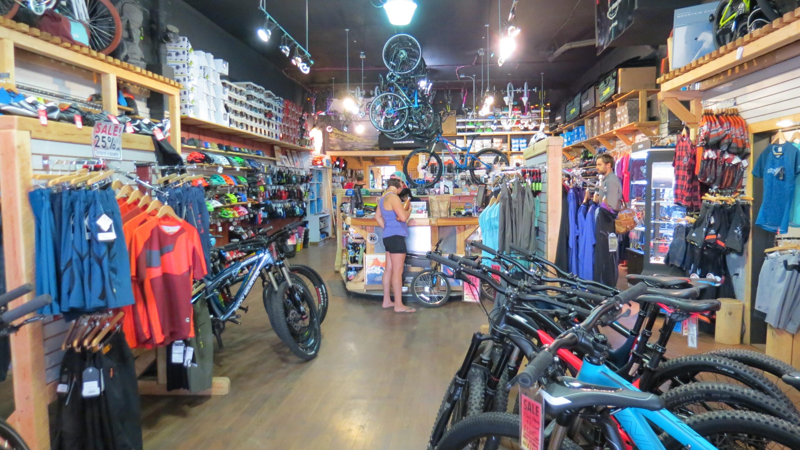 An indoor bike shop featuring a variety of bicycles and cycling gear. The space is well-lit, showcasing rows of helmets, clothing, and accessories. A woman stands at the counter, while a man browses nearby. Several bikes are displayed prominently, and there are signs indicating ongoing sales.