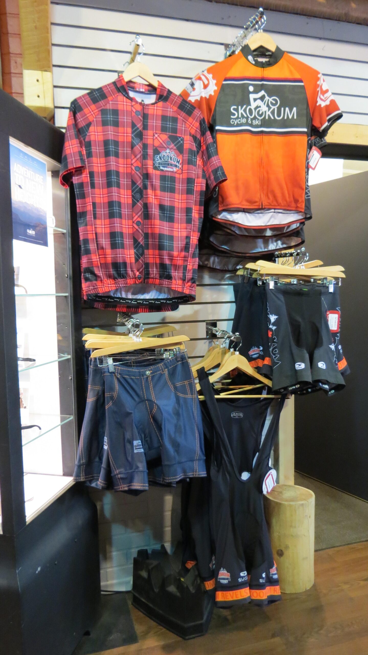 Display of cycling apparel, including a red plaid jersey and an orange cycling jersey, hanging on wooden hangers. Below are various styles of cycling shorts, including denim-like fabric and black options, arranged attractively in a retail setting.