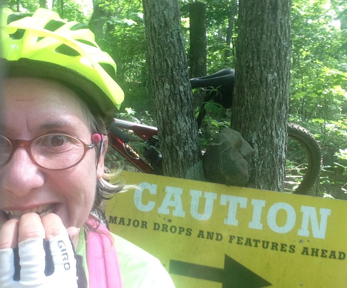 A person wearing a bright yellow helmet and cycling gloves smiles with excitement while holding their hands up near their face. In the background, there is a caution sign reading "Caution: Major Drops and Features Ahead," along with a red mountain bike partially visible among the trees in a lush, green forest setting. Rocky Knob Park mountain bike trail.