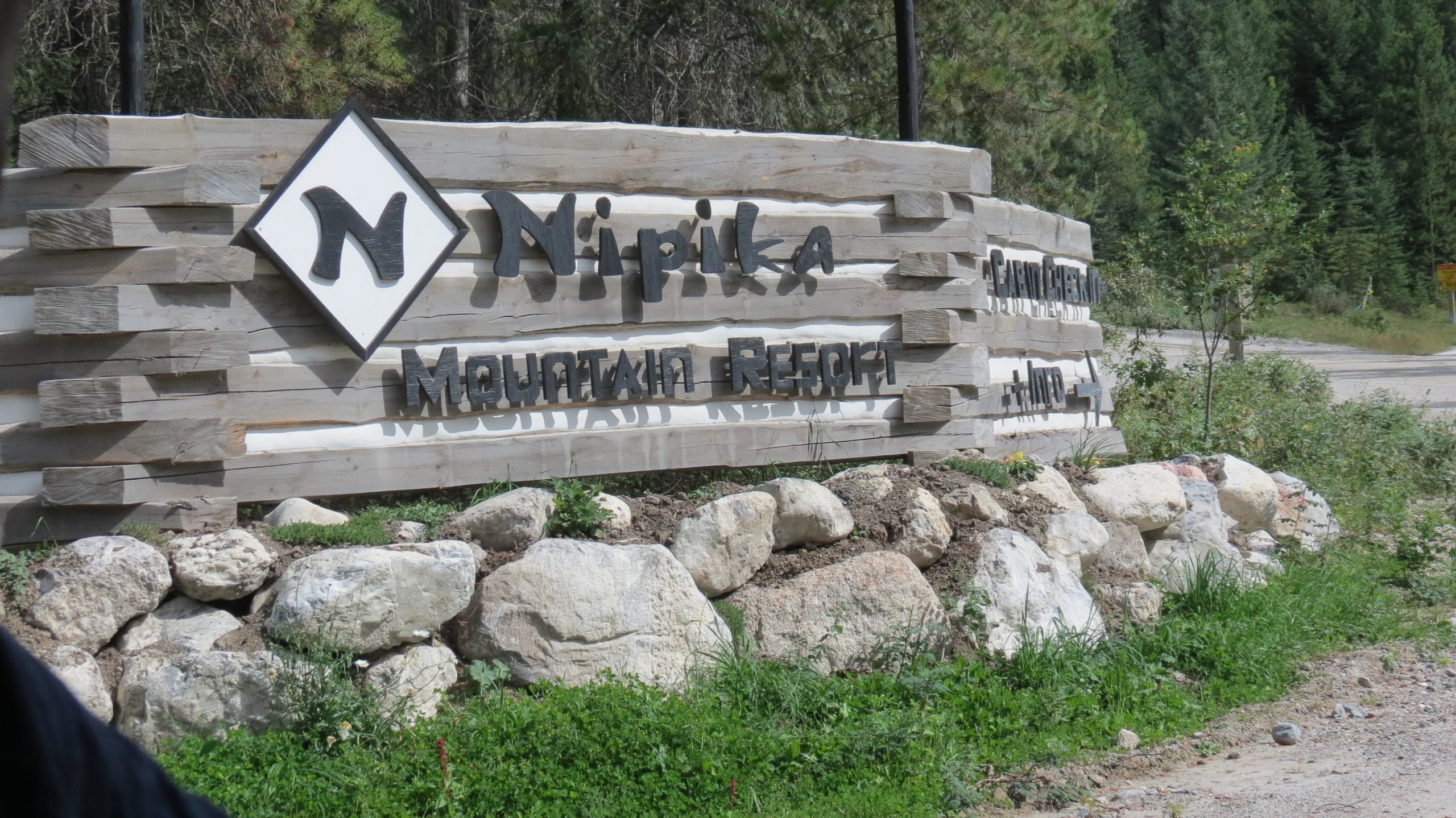 Sign for Nipika Mountain Resort, featuring wooden logs and stone landscaping, surrounded by greenery and trees. Nipika Mountain Resort mountain bike trail.