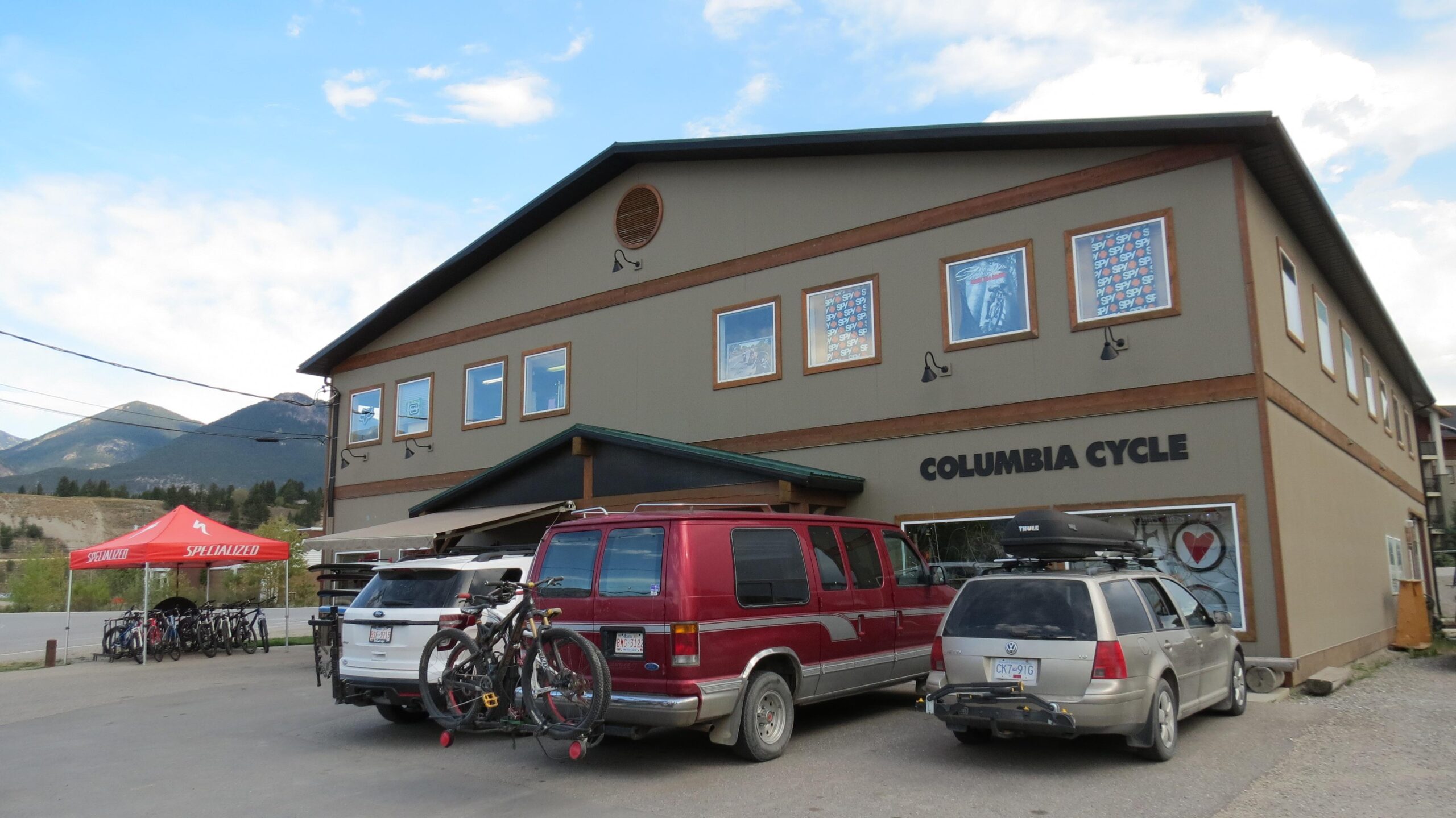 Exterior of Columbia Cycle, a bicycle shop, featuring a two-story building with large windows and a sign. Two vehicles, including a red van and a silver minivan, are parked out front, both equipped with bike racks. A red canopy with the Specialized logo is set up nearby, showcasing bikes for sale. The backdrop includes mountains under a clear blue sky.