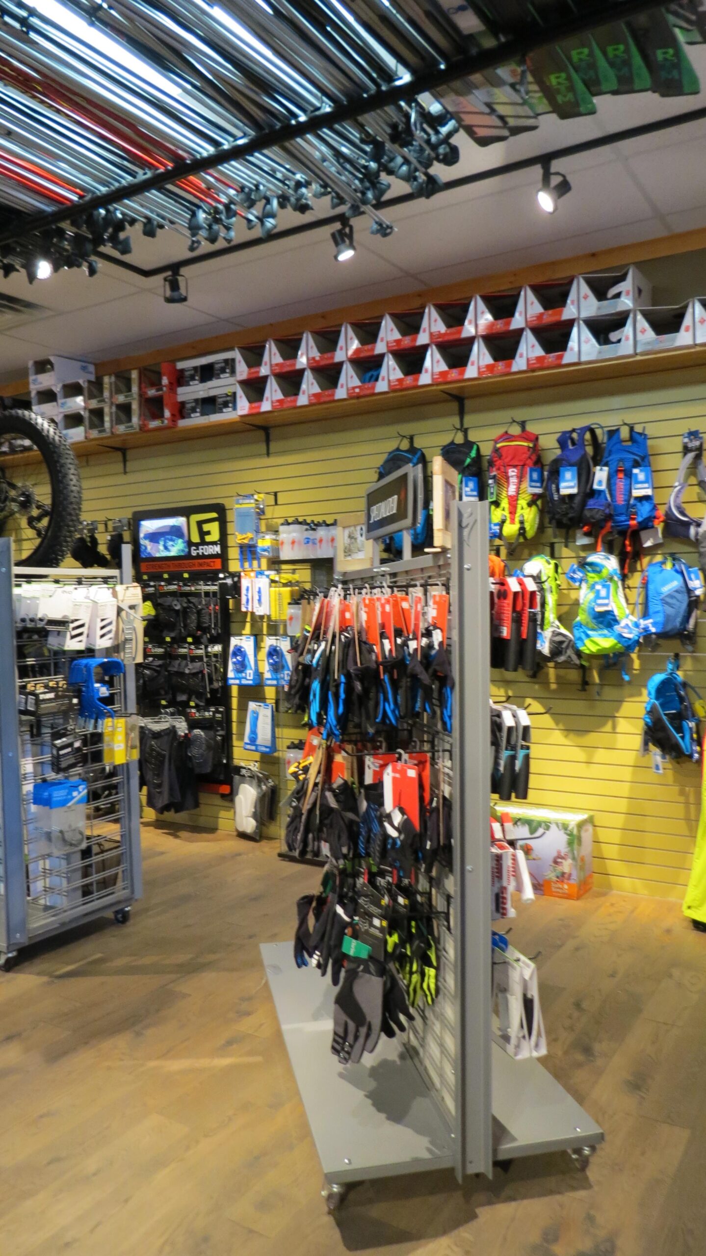 Inside a sporting goods store, various outdoor gear and equipment are displayed. The walls showcase a range of colorful backpacks, gloves, and protective gear, while shelves above hold neatly arranged boxes. The ceiling features metal poles adding structure, and bright lighting highlights the merchandise, creating an inviting atmosphere for shoppers interested in outdoor activities.