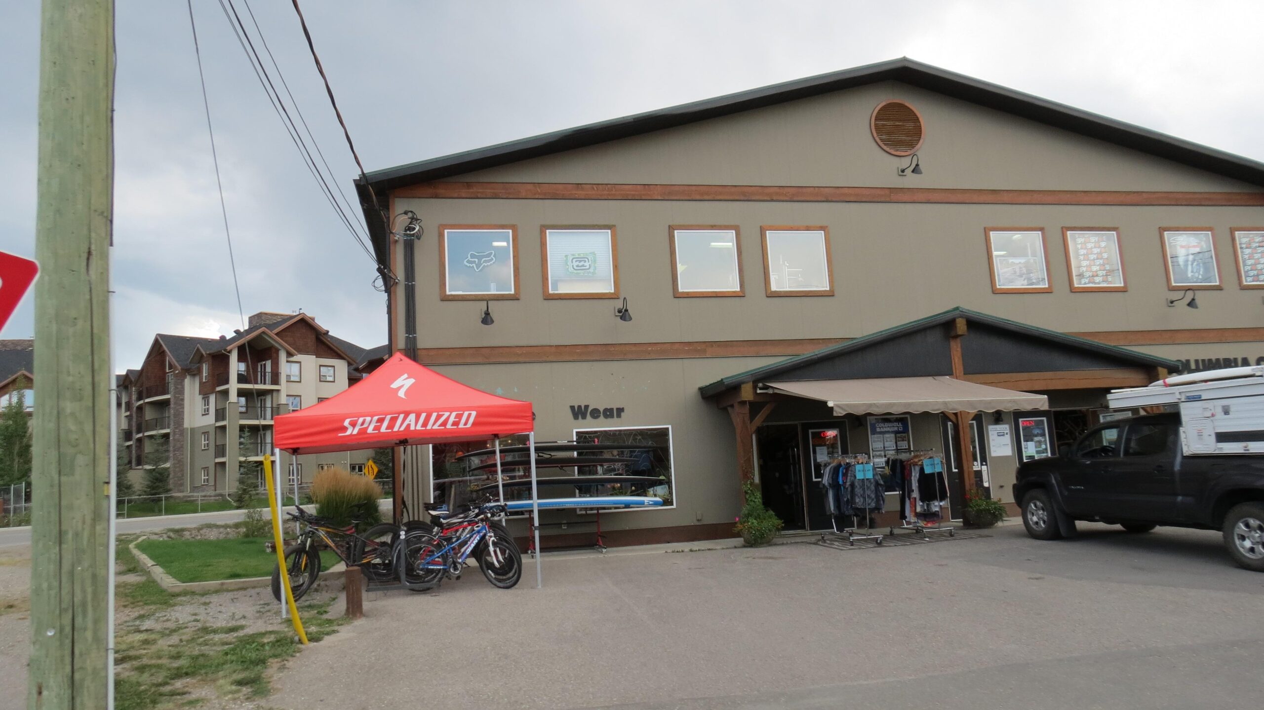 A storefront of a sporting goods shop featuring outdoor clothing and equipment, with a red canopy displaying the Specialized logo. Bicycles are parked outside, and there are displays of paddleboards visible in the windows. Surrounding buildings are partially visible in the background under a cloudy sky.