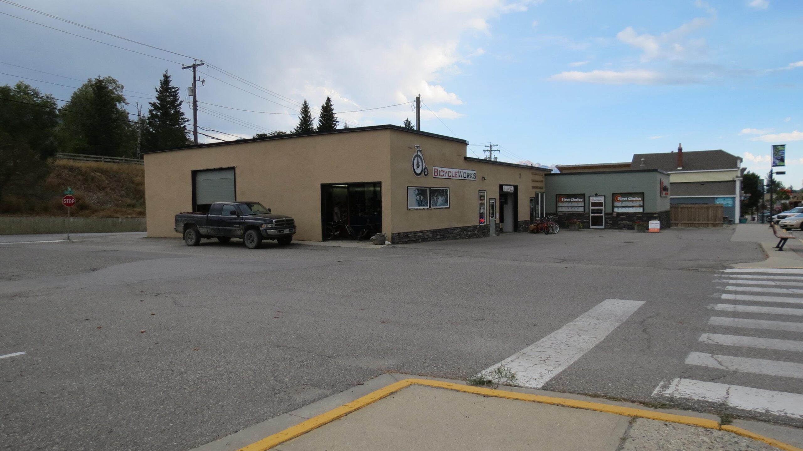 A view of a bicycle shop called "Bicycle Works," featuring a tan building with large windows and a sign. In front, a dark pickup truck is parked on a paved lot. Nearby, there are additional buildings and a stop sign, indicating a quiet street setting with trees in the background and partly cloudy skies above.