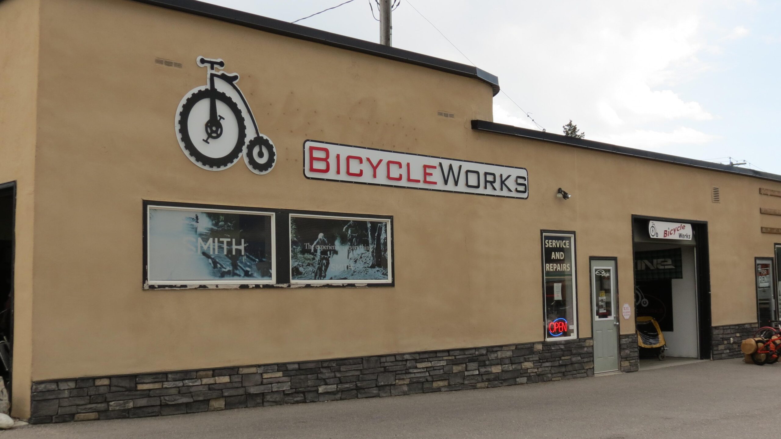 Exterior view of a bicycle shop named "Bicycle Works," featuring a bike logo on the wall, multiple windows, and a sign that reads "SERVICE AND REPAIRS." The entrance shows an "OPEN" neon sign. The building has a beige stucco finish and a stone base.