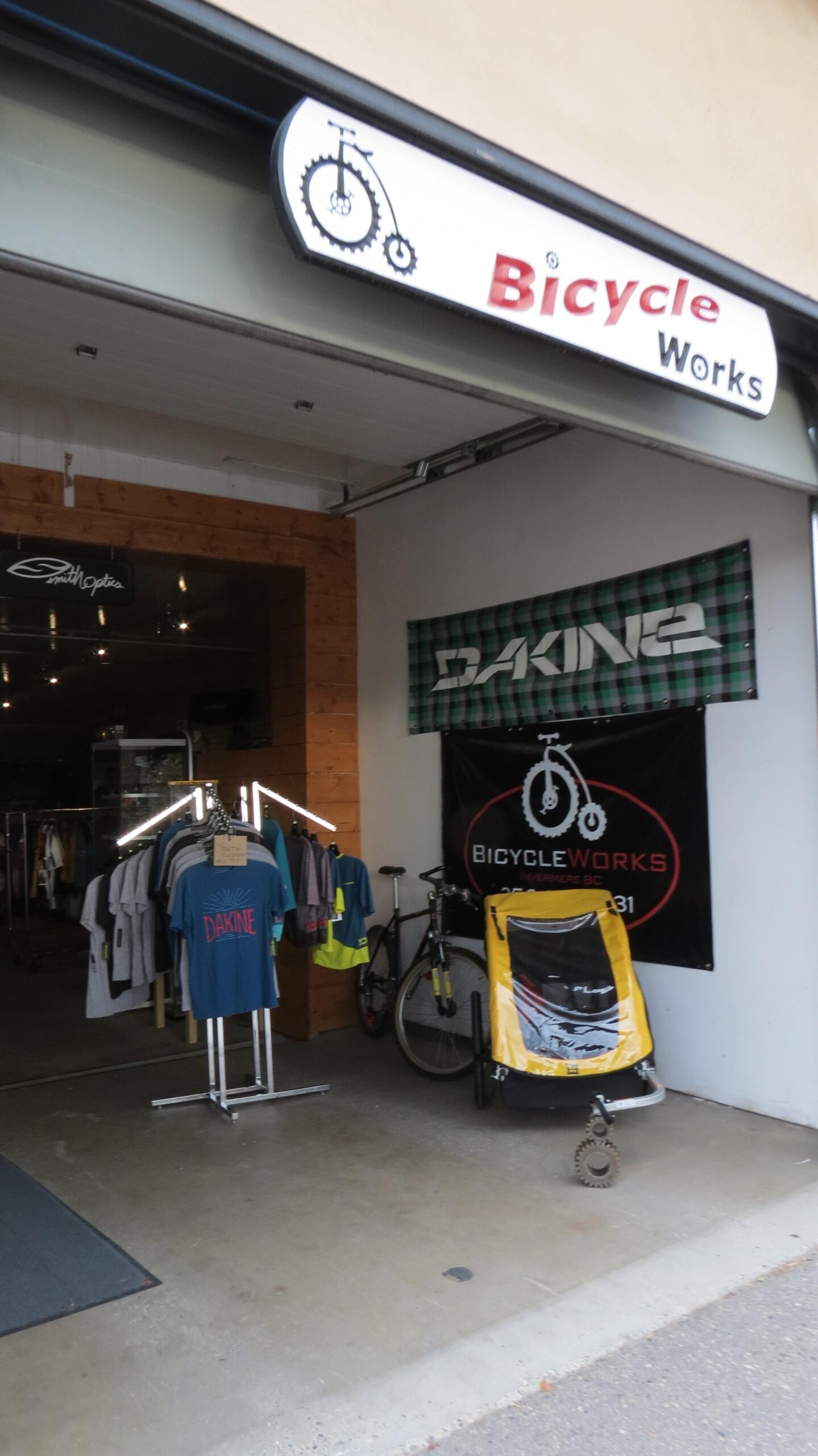 Entrance of a bicycle shop called "Bicycle Works," featuring a sign with a bicycle logo. The storefront displays clothing on racks, including shirts with the brand "Dakine." There is also a bicycle and a yellow bike trailer visible inside the shop. The interior has wooden paneling and banners promoting various brands.
