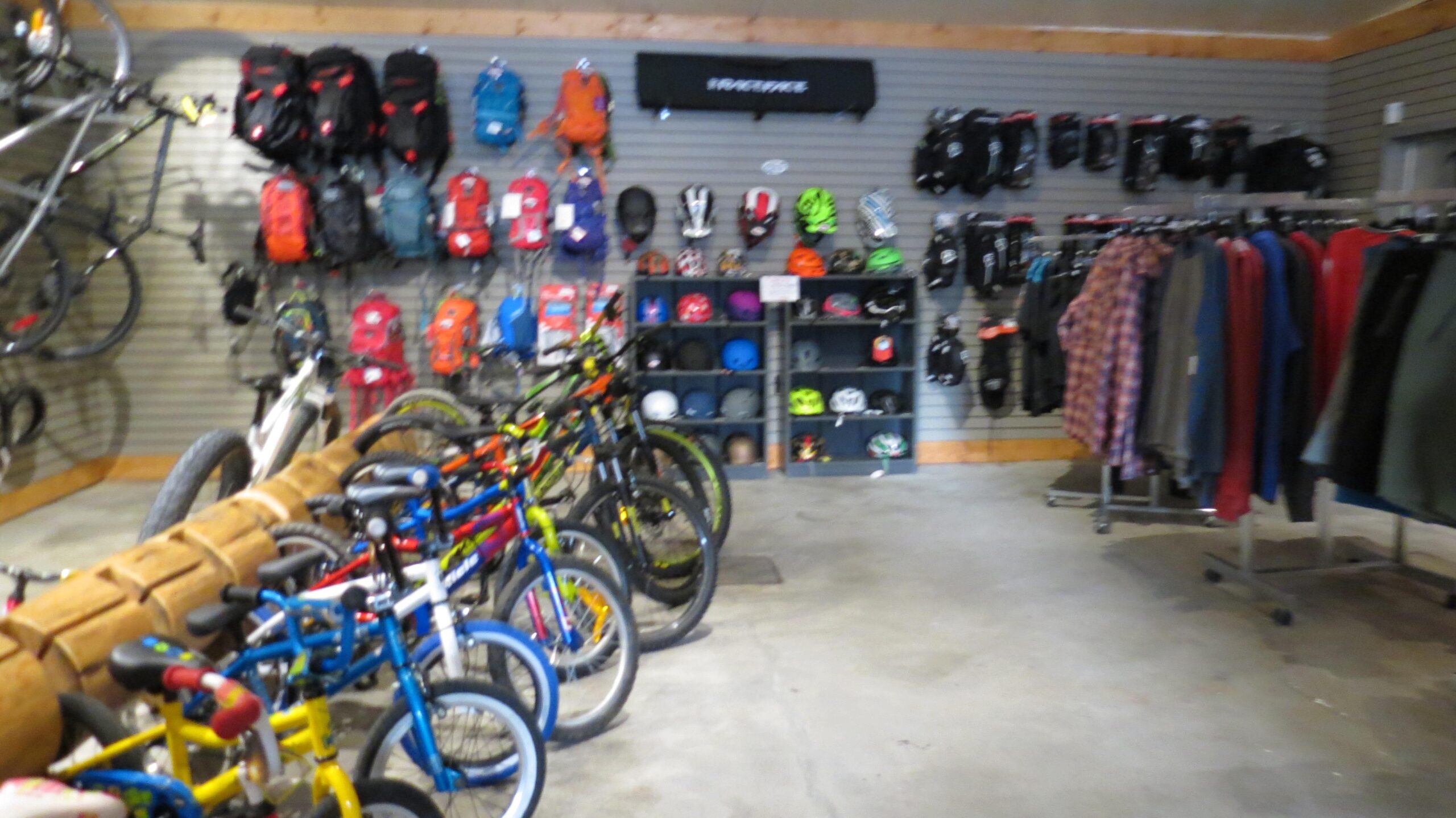 A view of an indoor shop showcasing a variety of bicycles, including children's bikes in bright colors, along with a display of backpacks and helmets on the walls. Clothing items are neatly hung on racks, and the space has a concrete floor and wooden accents.