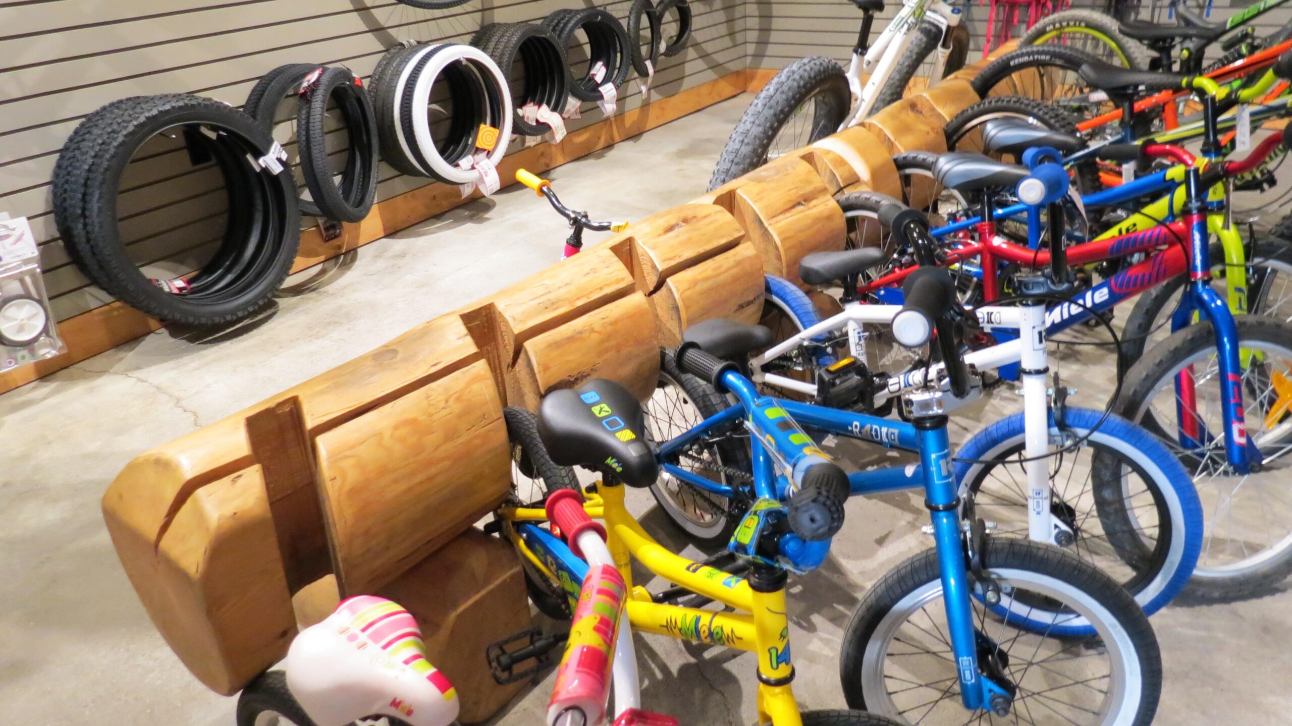 A display inside a bicycle shop featuring several colorful kids' bicycles parked next to a large wooden log display. In the background, various bicycle tires hang on the wall. The floor is concrete, and the shop has an organized, inviting atmosphere.