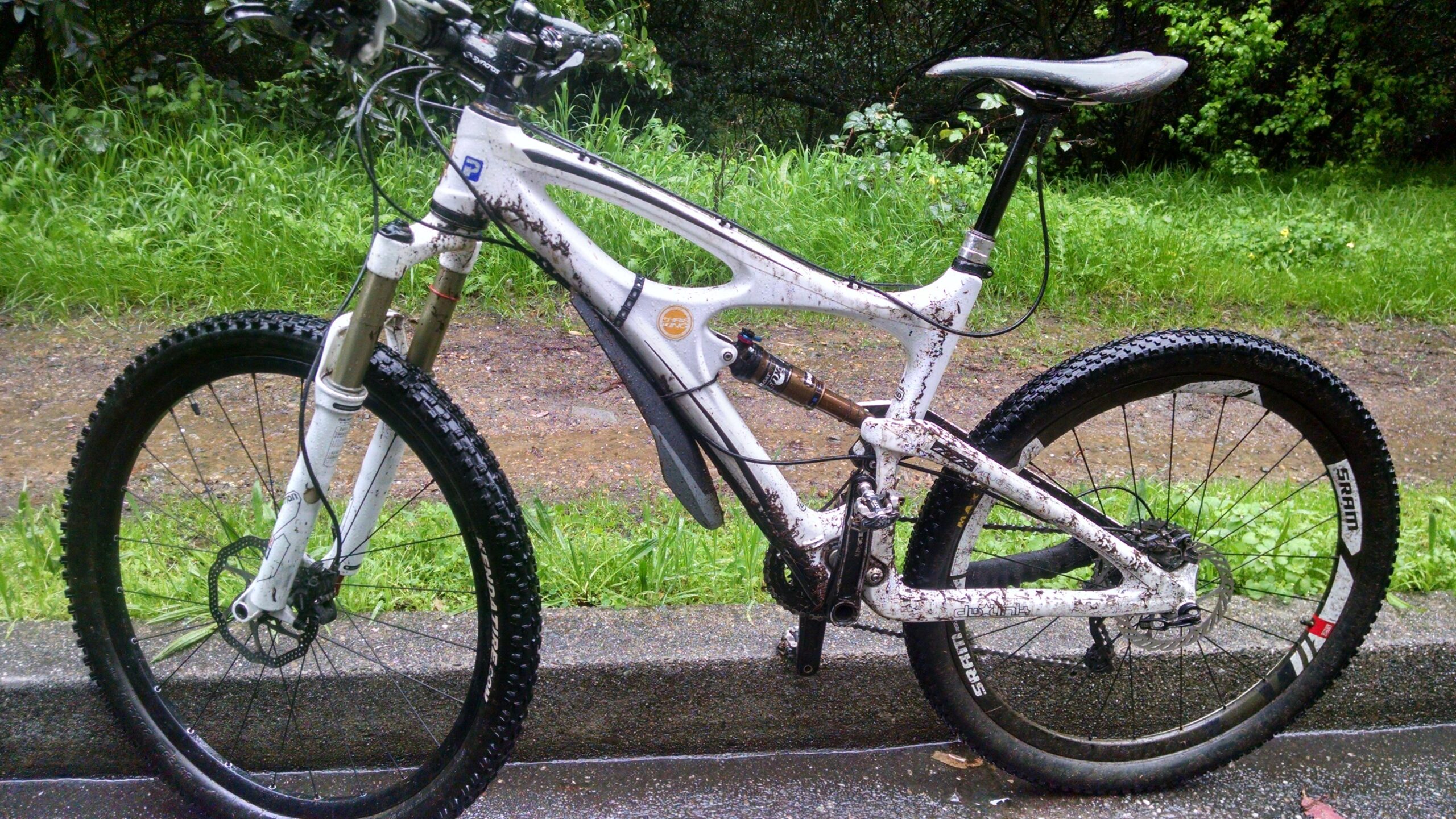 Ibis Mojo SL-R: A muddy mountain bike partially leaning against a concrete edge, with a grassy area and a dirt path in the background. The bike features a white frame with visible dirt and mud splatters, showcasing its use in outdoor terrain. The wheels are equipped with thick, knobby tires designed for off-road riding.