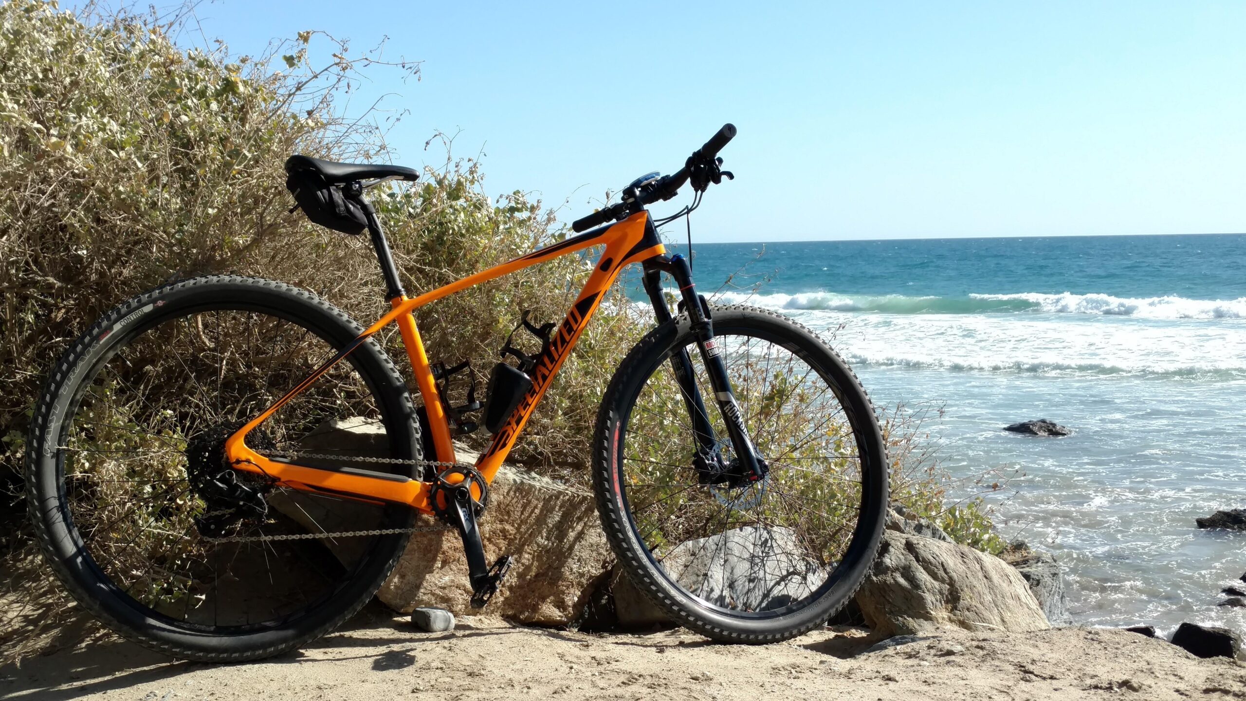 Specialized Stumpjumper: A vibrant orange mountain bike is parked on a sandy beach next to the ocean, with gentle waves rolling in the background and bushes lining the shore under a clear blue sky.