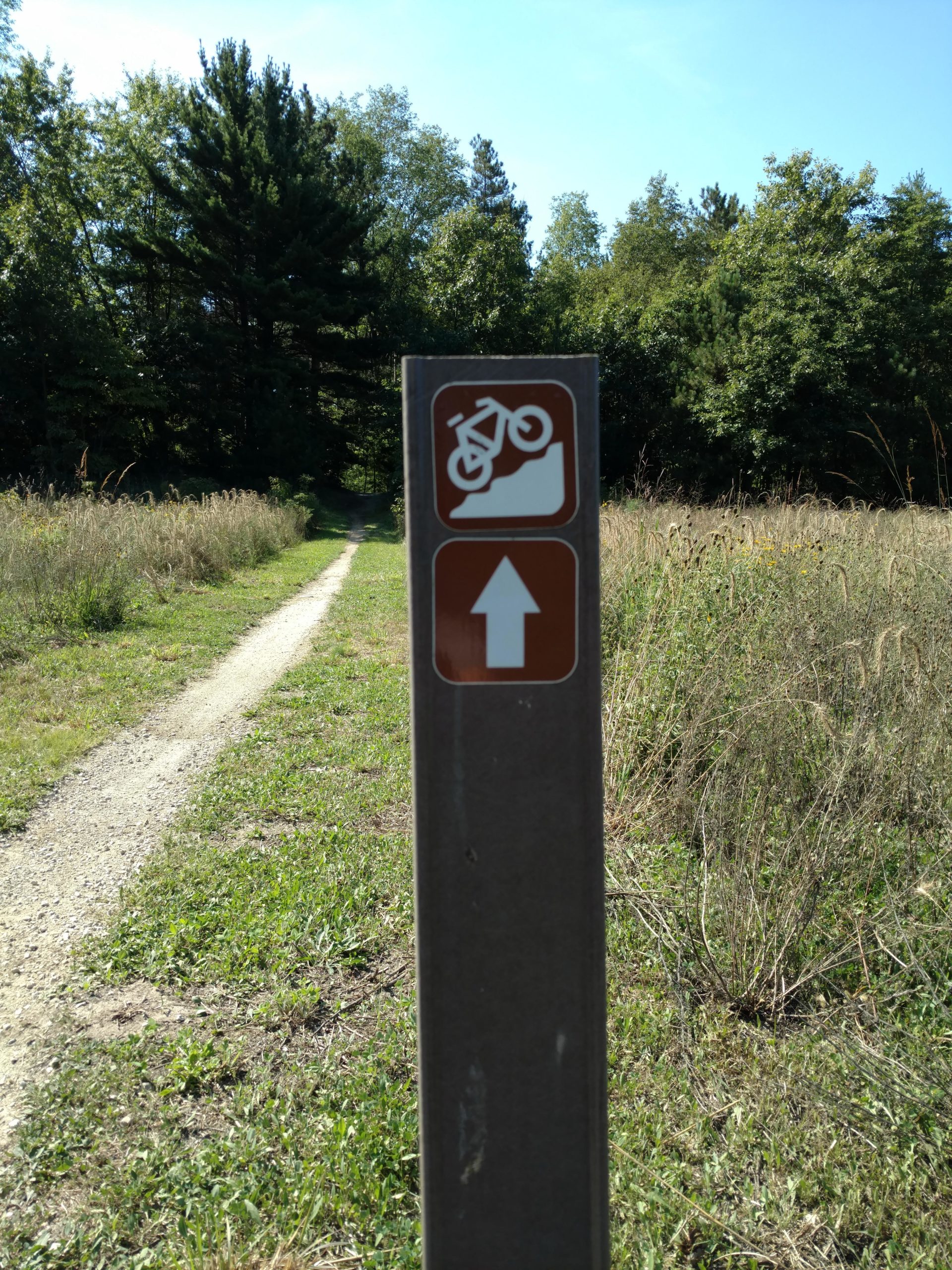 Sign marking a mountainous biking trail, featuring an icon of a bike going uphill and an upward arrow. The sign is positioned along a dirt path surrounded by grass and trees under a clear blue sky. Riley Trails mountain bike trail.