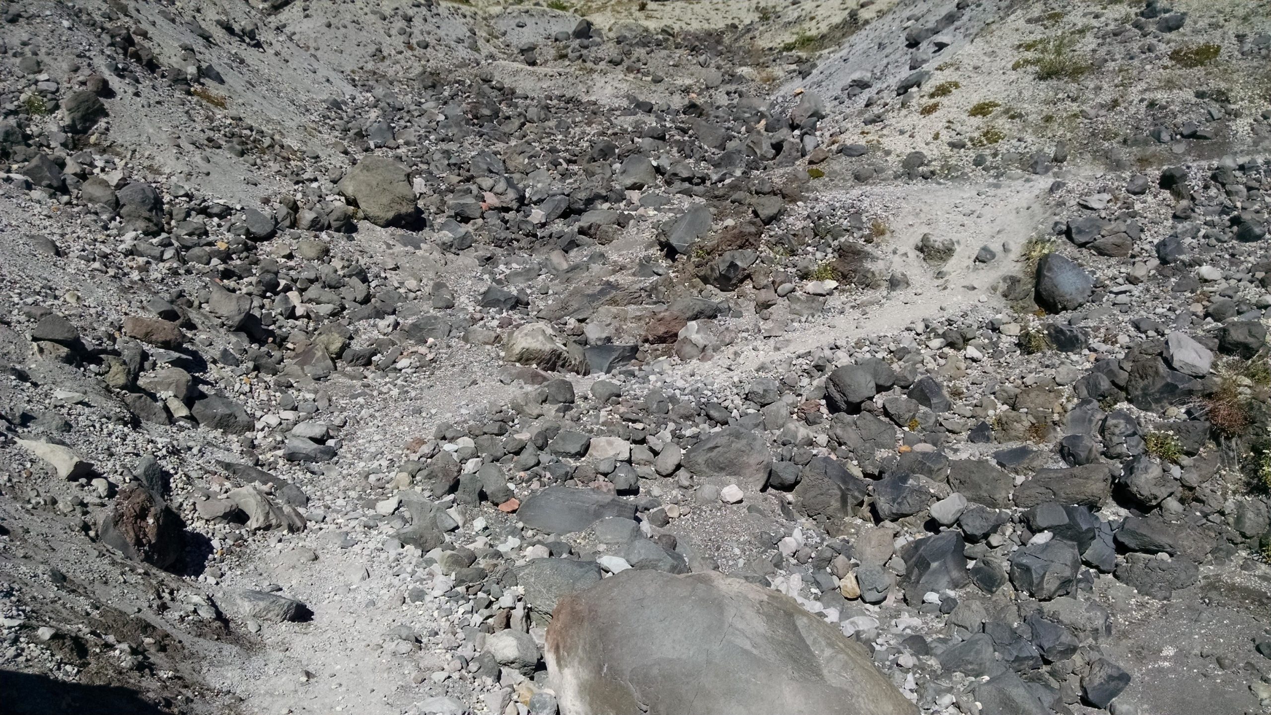 A rocky, volcanic landscape featuring a variety of sizes of stones and gravel scattered across a dry, sandy surface. The terrain includes both larger boulders and smaller pebbles, emphasizing the rugged and uneven topography. Sparse vegetation can be seen in patches, highlighting the arid conditions of the area. Ape Canyon#234, Abraham#216d, Smith Creek#225 Trails mountain bike trail.