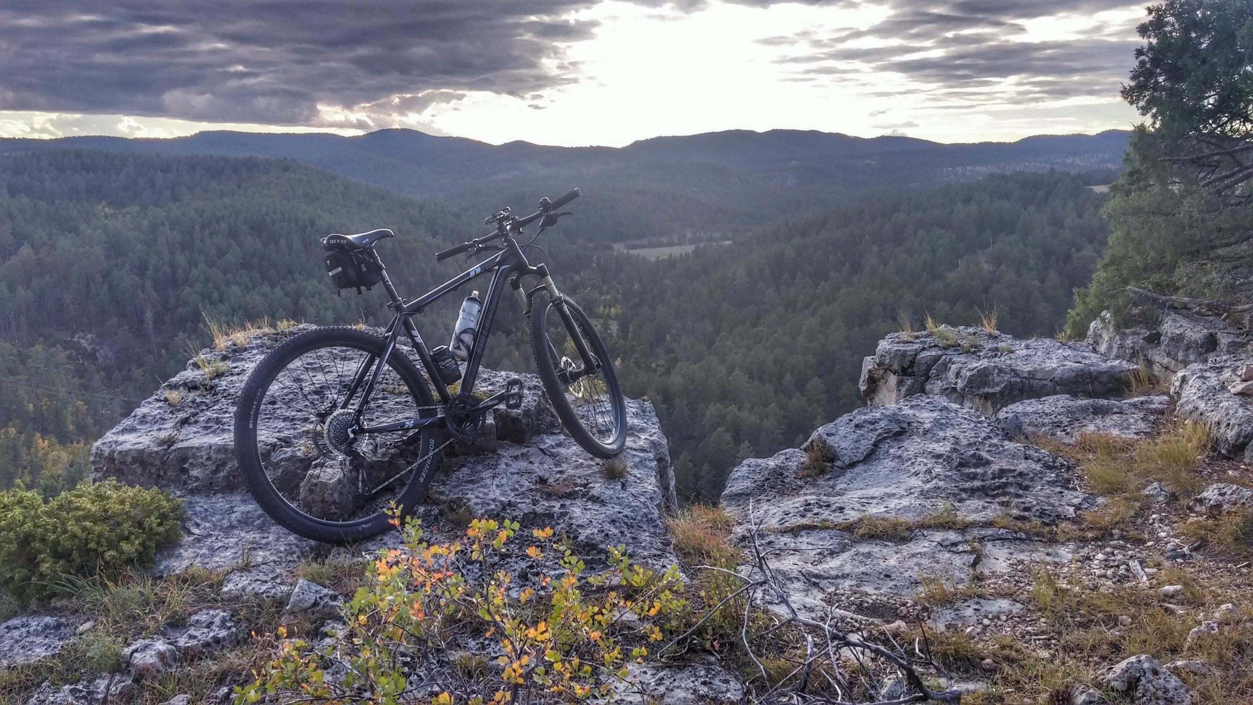 A mountain bike resting on a rocky ledge, with a breathtaking view of a lush green valley and distant mountains under a cloudy sky during sunset. Victoria's Secret mountain bike trail.