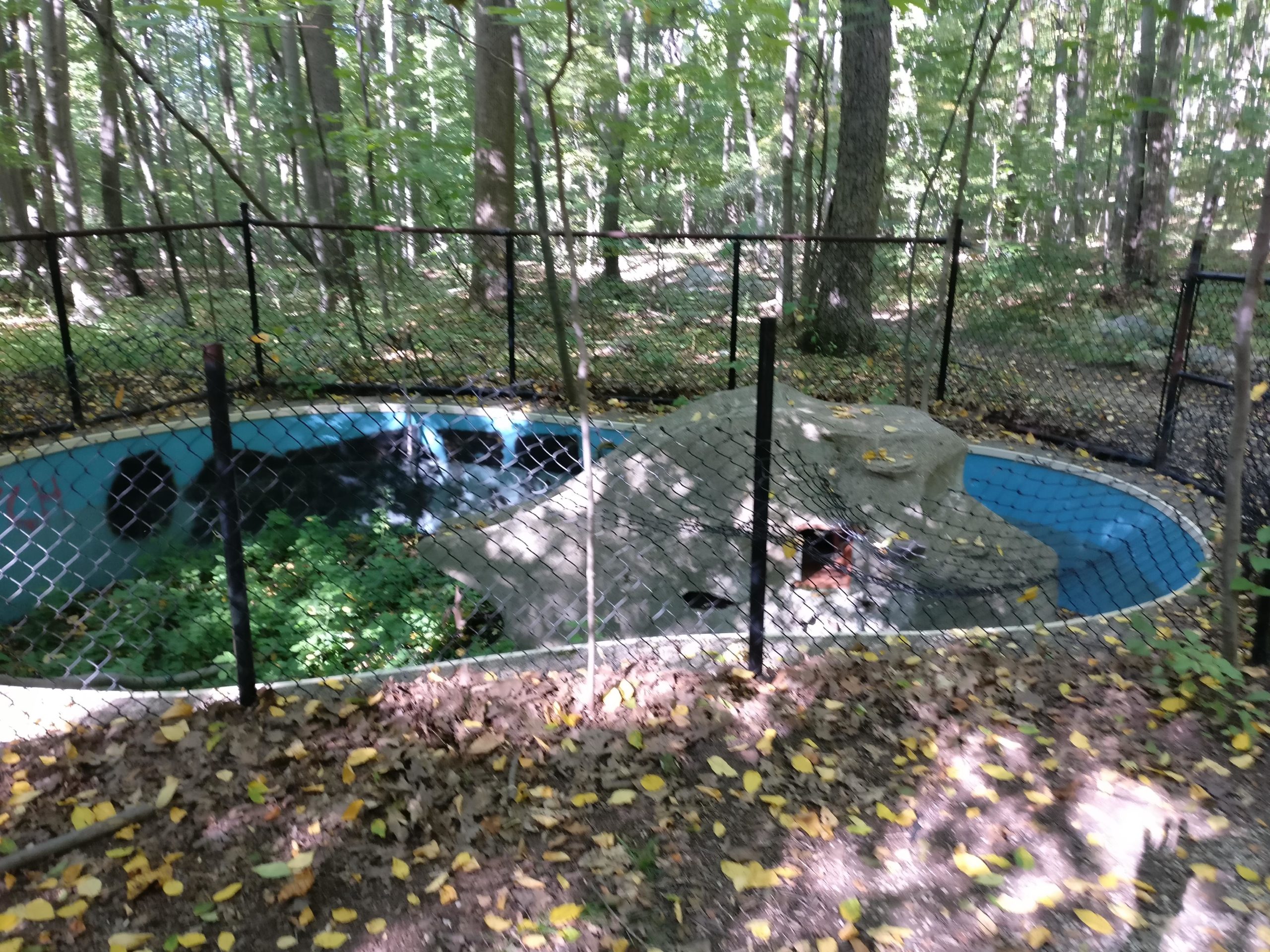 A dilapidated circular pool surrounded by a black chain-link fence, situated in a wooded area with green foliage and scattered yellow leaves on the ground. The pool has a blue interior and a rocky structure on one side, indicating disuse. Sunlight filters through the trees, casting dappled light on the scene. Long Pond Ironworks State Park mountain bike trail.