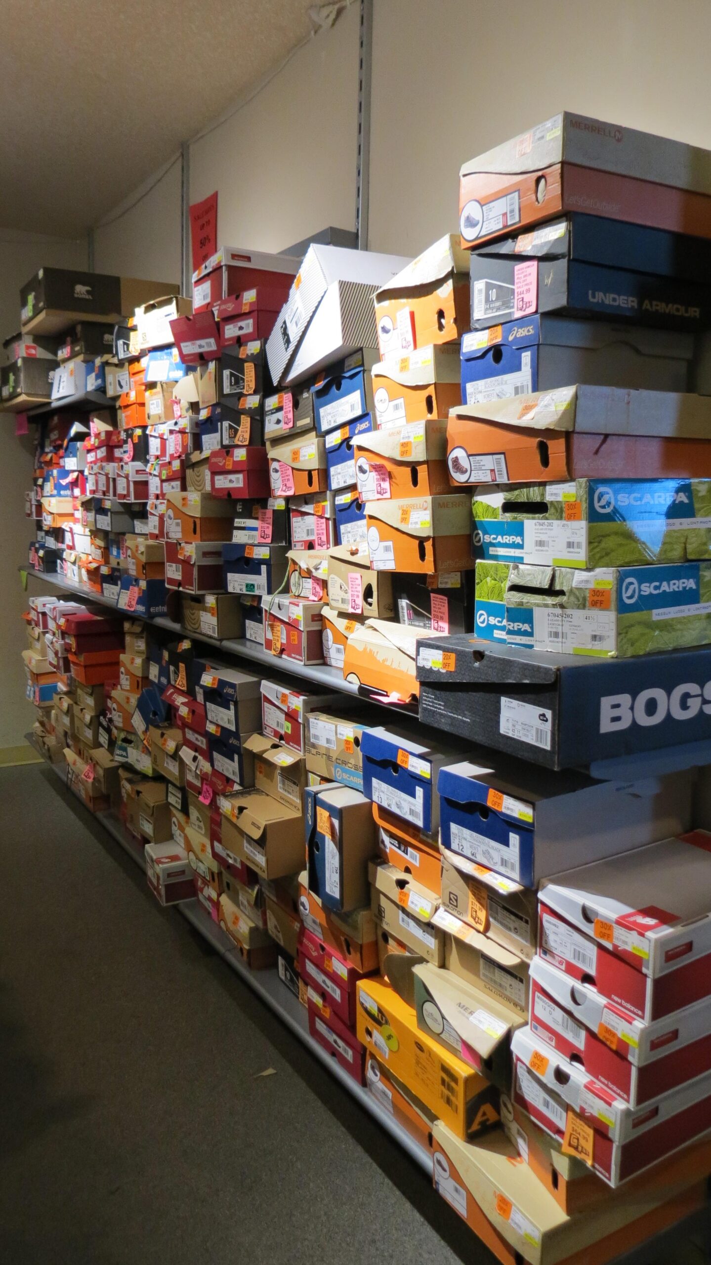 A cluttered display of various shoe boxes stacked on shelves, with many boxes featuring colorful labels and price tags. The shelves extend across a store interior, showcasing a wide selection of shoe brands.