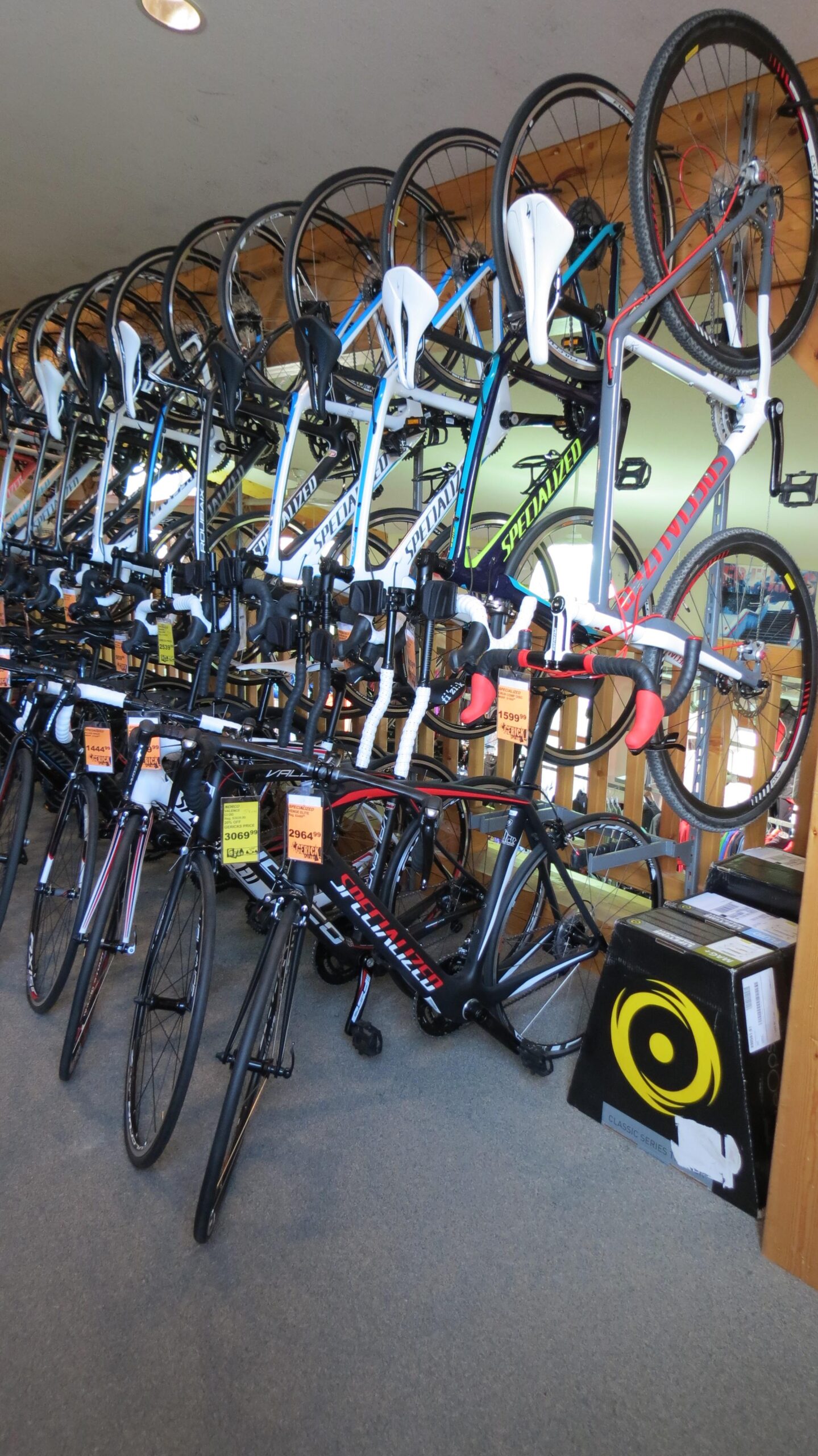 A display of various road bicycles arranged on racks in a bike shop. The bikes feature different colors and brands, including several with "Specialized" branding. Price tags are visible on some of the bicycles, and a few boxes are positioned on the floor in the background. The interior has wooden beams and a well-lit atmosphere.