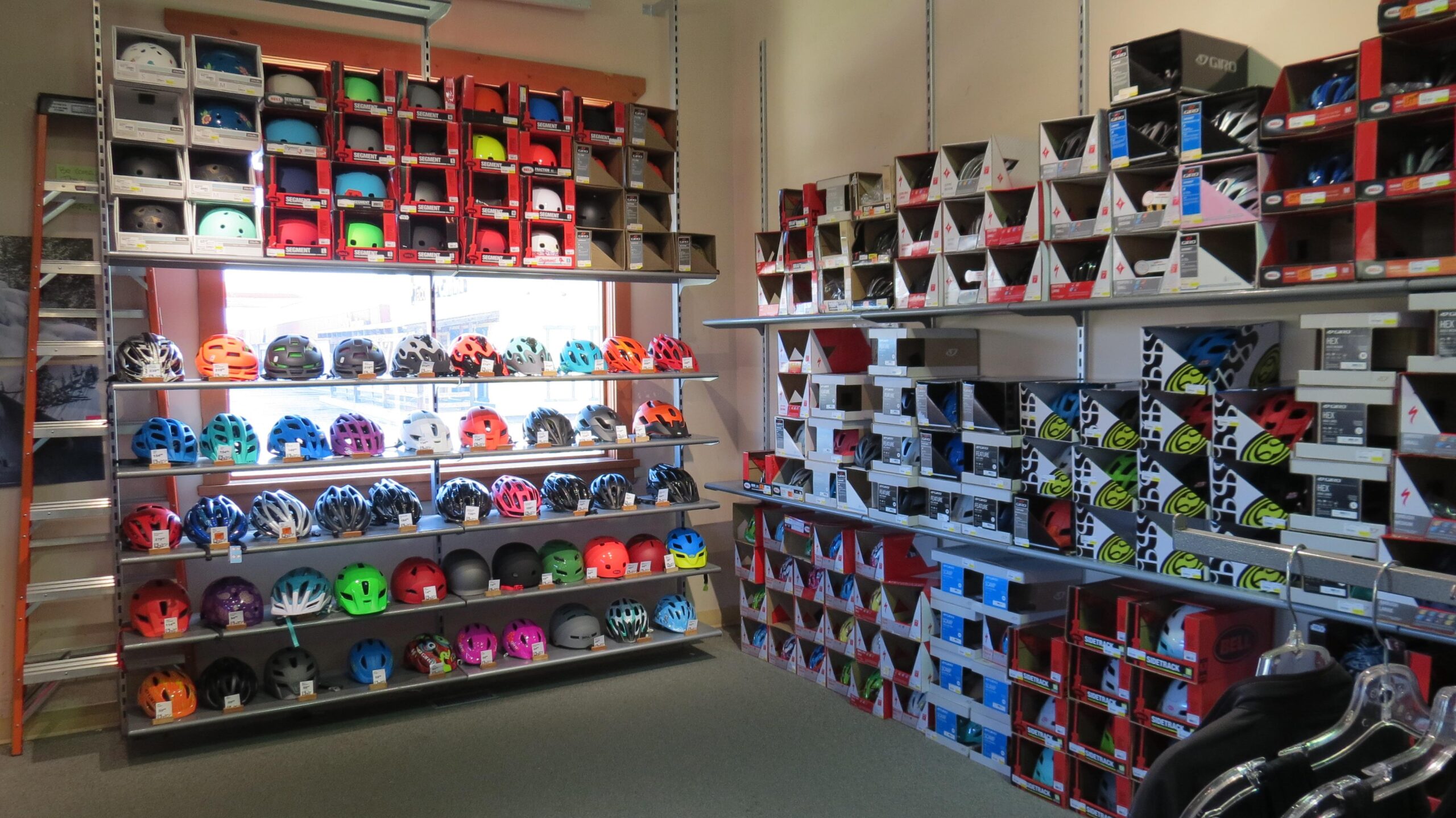 A retail display of various bicycle helmets arranged on shelves, showcasing a wide range of colors and styles. The scene features multiple rows of helmets, with some models displayed in their original packaging. An orange ladder is visible on the left, and a window in the background lets in natural light.