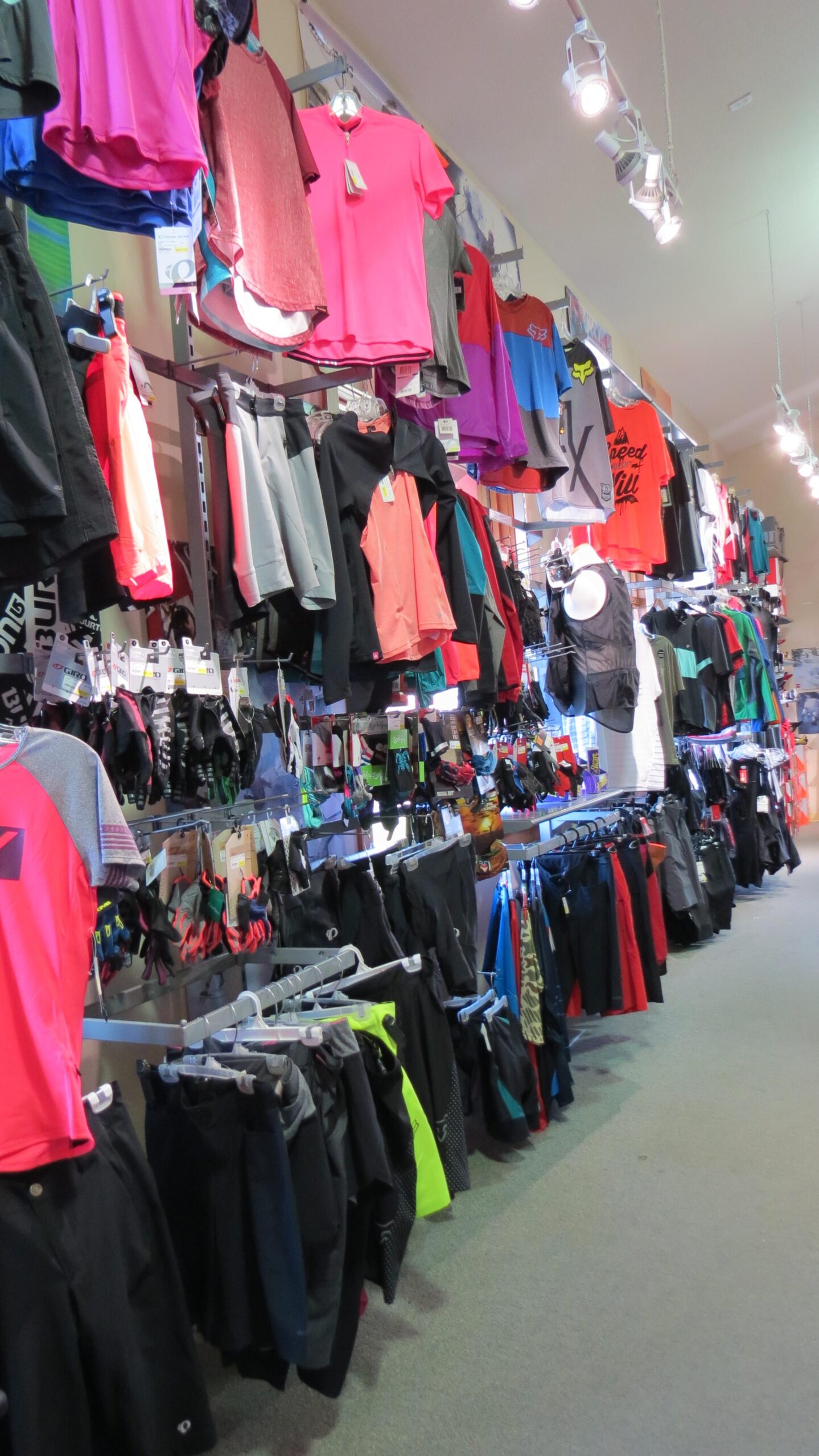 A retail display featuring a variety of athletic clothing, including T-shirts, shorts, and gloves, organized on racks. The clothes are in various colors such as pink, red, black, blue, and green, with tags still attached. The background shows bright lighting and a neatly arranged store interior.