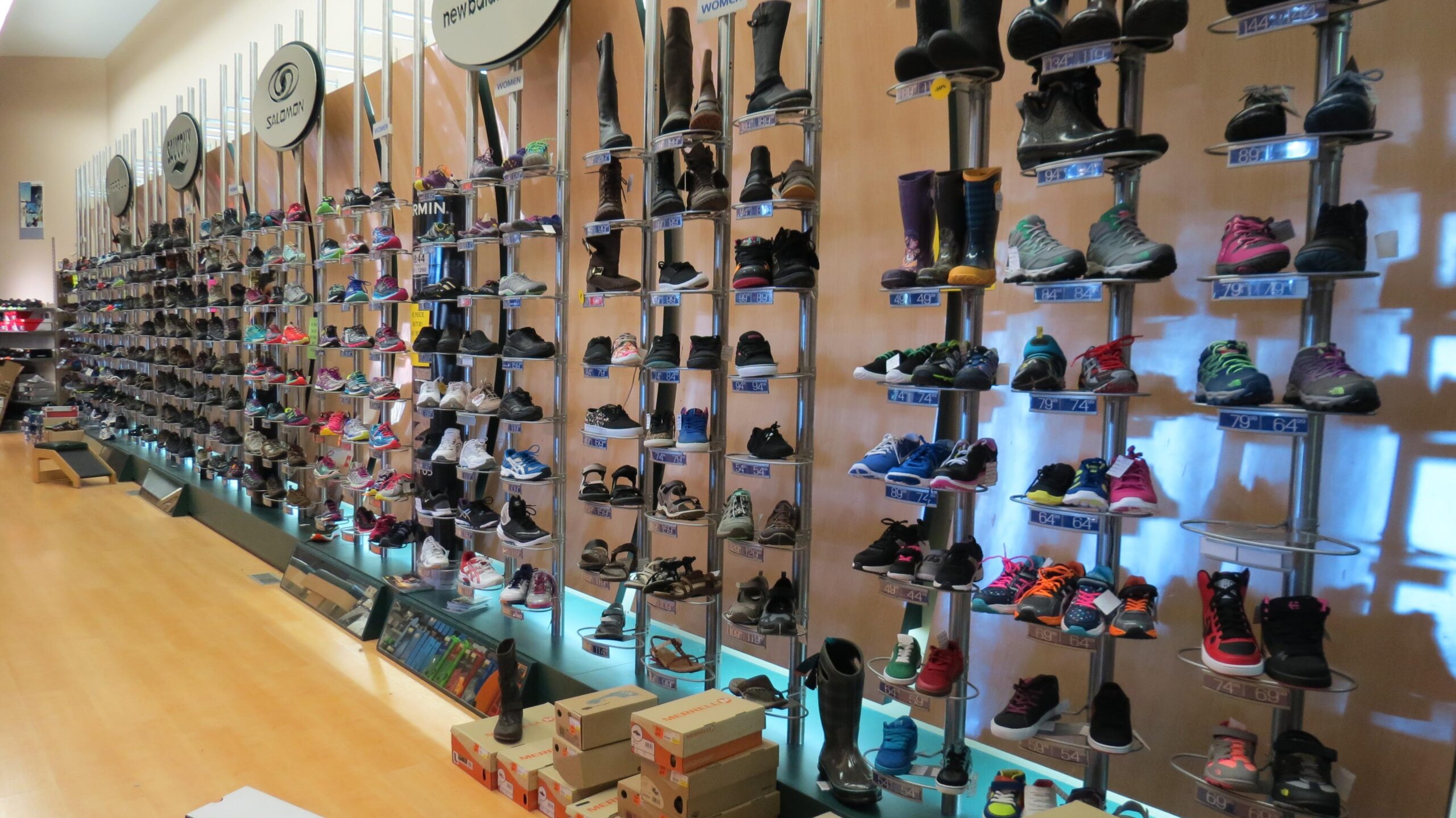 A neatly arranged display of various types of shoes on multiple shelves in a sporting goods store. The collection includes athletic shoes, boots, and sandals in different colors and styles, with pricing information visible on labels. The store features wooden flooring and bright lighting that enhances the visibility of the shoes.