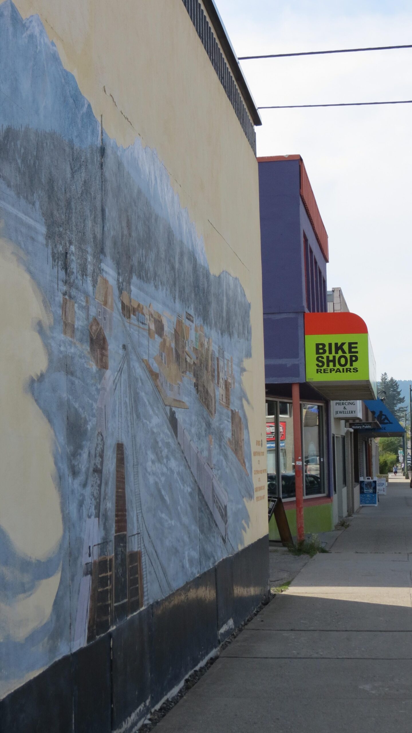 A view of a city street featuring a colorful bike shop sign on the right. On the left, a large mural depicting a landscape with mountains, trees, and a historical town scene extends along the wall. The sidewalk is visible, with some greenery and additional storefronts in the background.