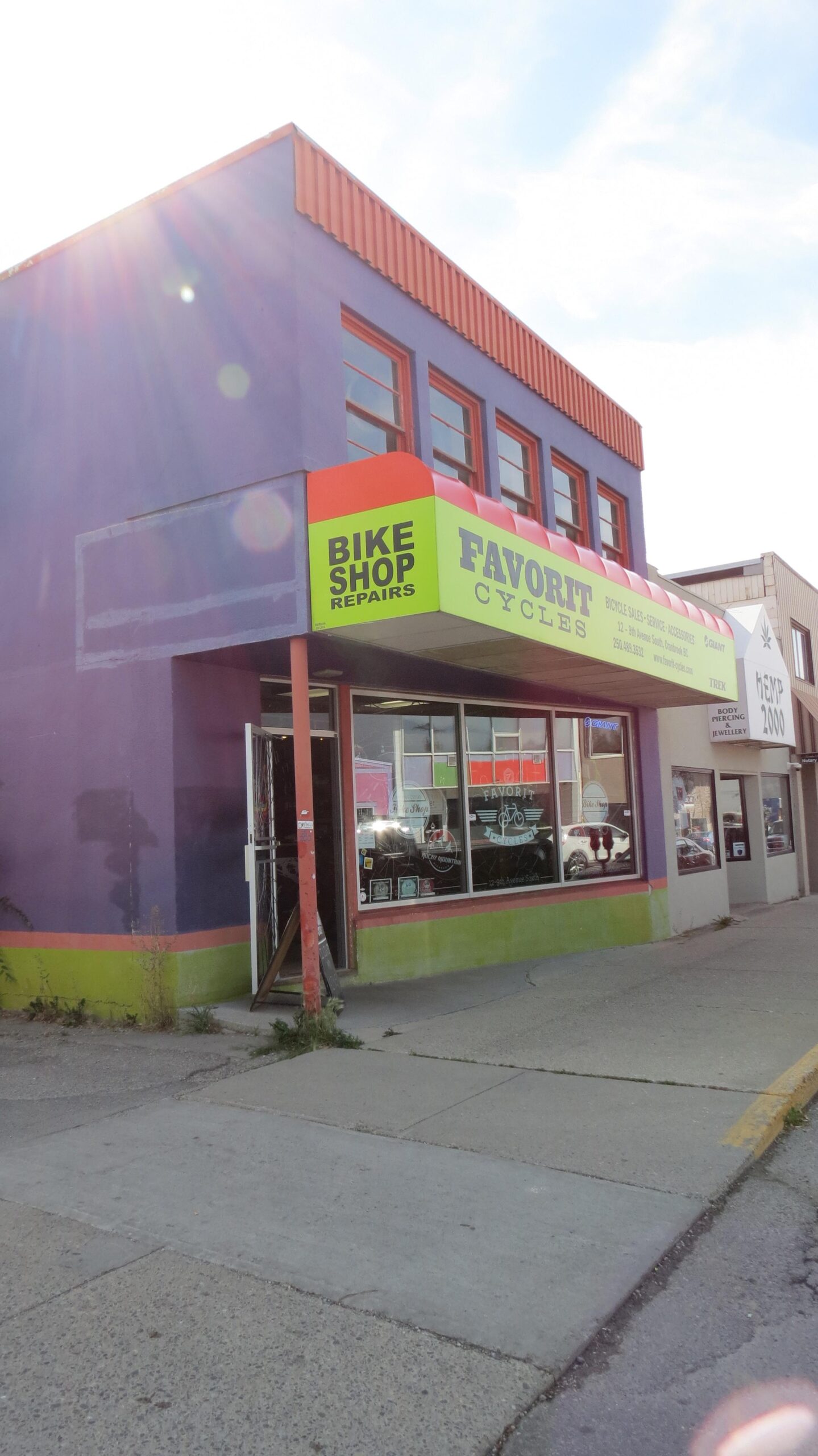 A colorful bike shop storefront featuring a purple exterior with bright green and orange accents. The sign reads "Favorit Cycles" and "Bike Shop Repairs." The entrance shows an open door, and large windows display various bike accessories and equipment. The surrounding area includes a sidewalk and nearby buildings.