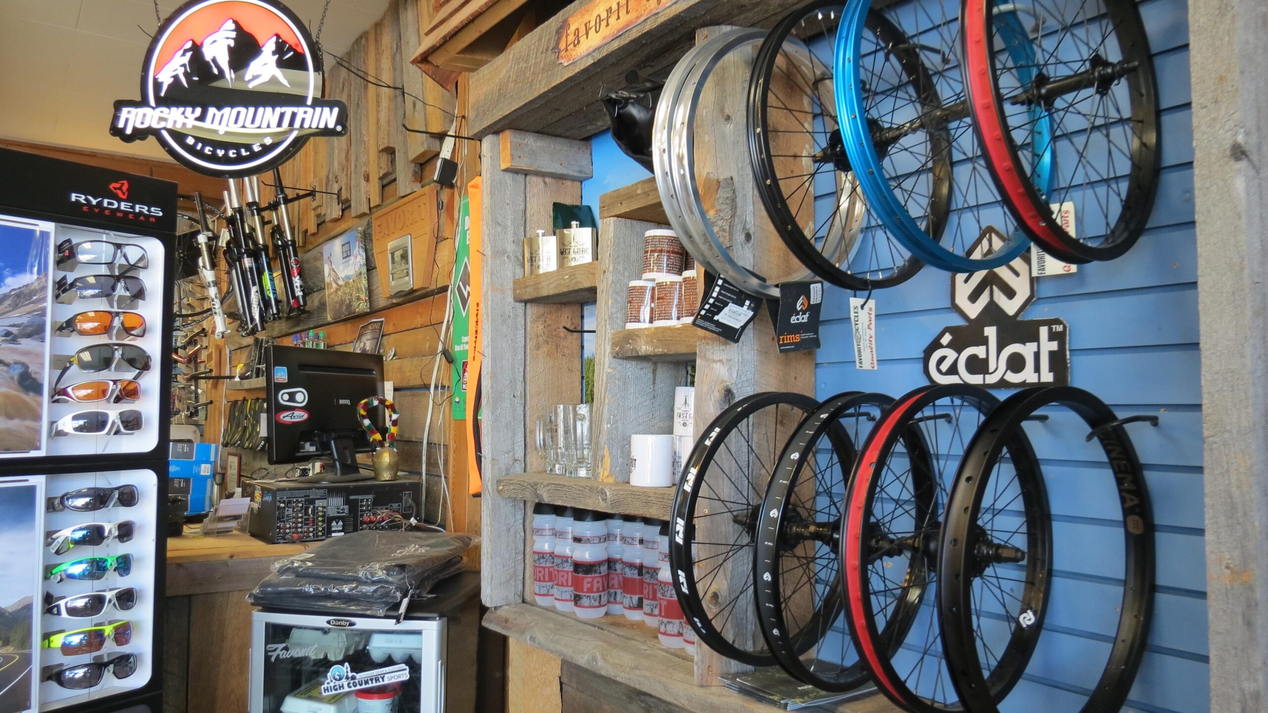 A display inside a bicycle shop featuring various bicycle wheels hanging on a wooden wall, alongside cycling accessories such as sunglasses, forks, and branded merchandise. The sign "Rocky Mountain Bicycles" is illuminated, and a checkout area with a computer and audio equipment is visible in the background.