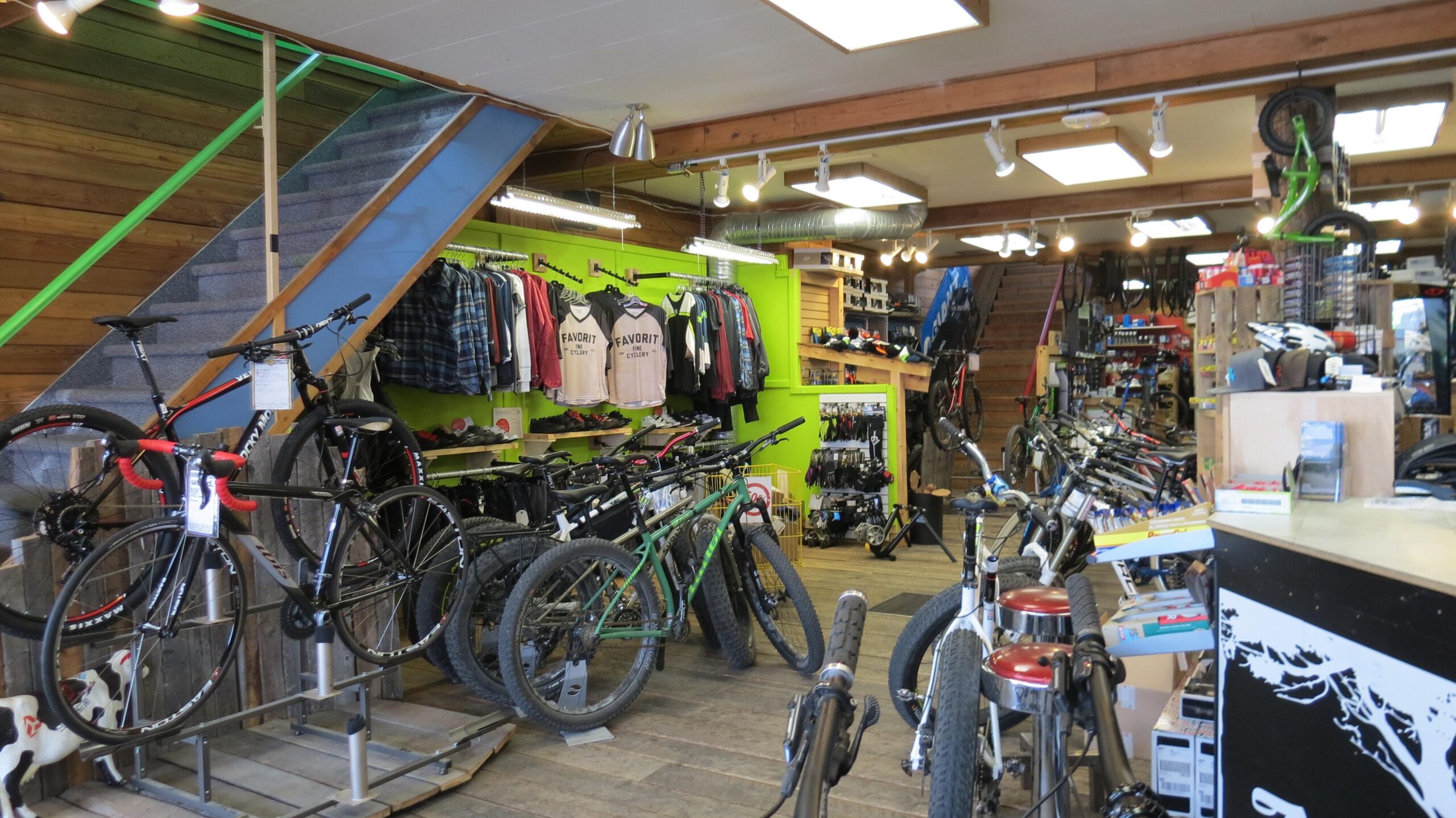 Inside a bike shop showcasing a variety of bicycles, including mountain bikes and road bikes, arranged throughout the space. The walls feature colorful displays of cycling apparel and accessories. A staircase leads to an upper level, and the shop has wooden flooring and bright lighting.