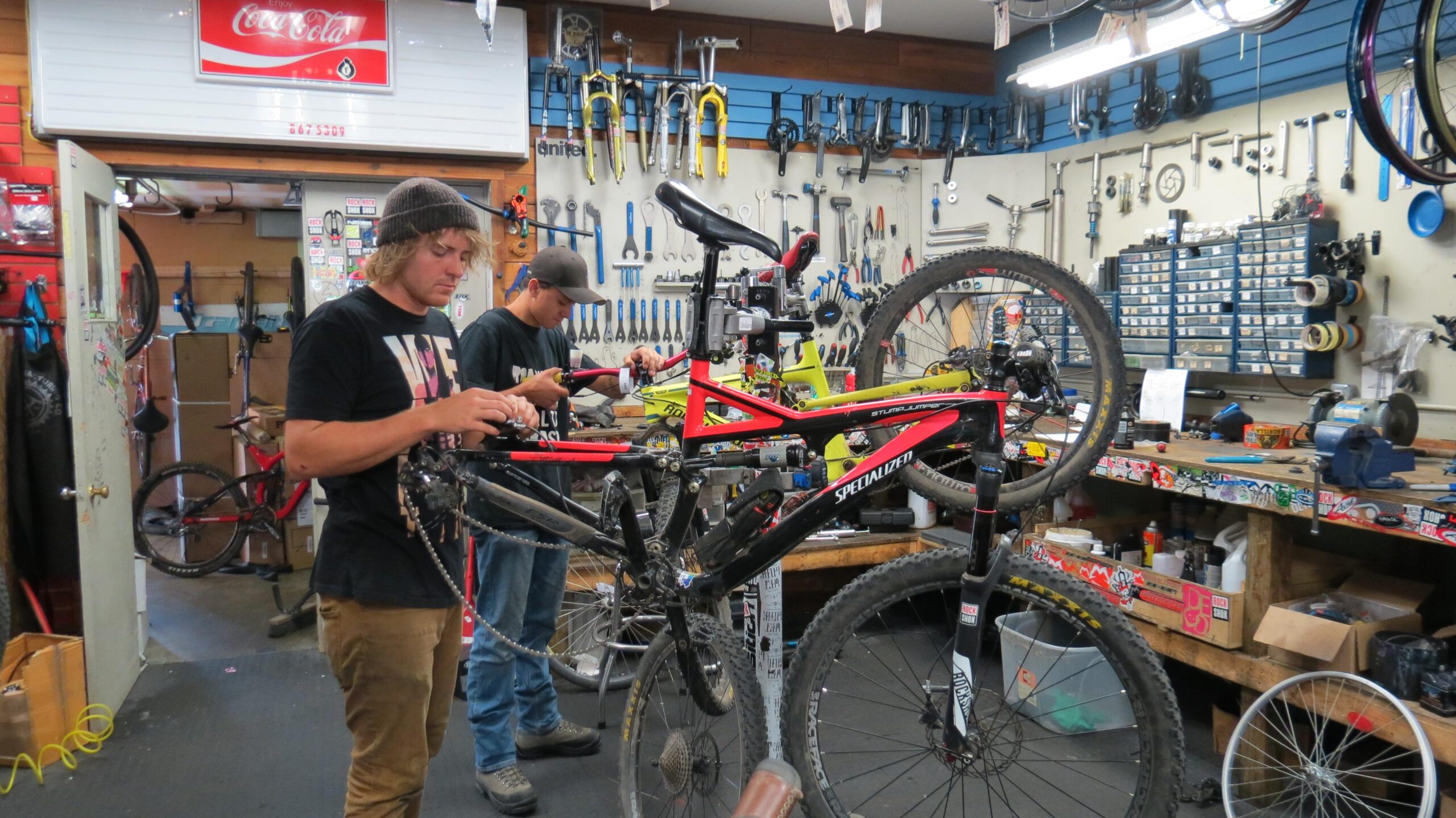Two mechanics are working on bicycles in a busy bike repair shop. One mechanic, in a black shirt and beanie, is adjusting the gears on a red and black mountain bike. The other mechanic, wearing a cap and black shirt, is repairing a different bike. The shop is filled with various bike tools and equipment, with bicycles hanging on the walls and workbenches cluttered with parts.