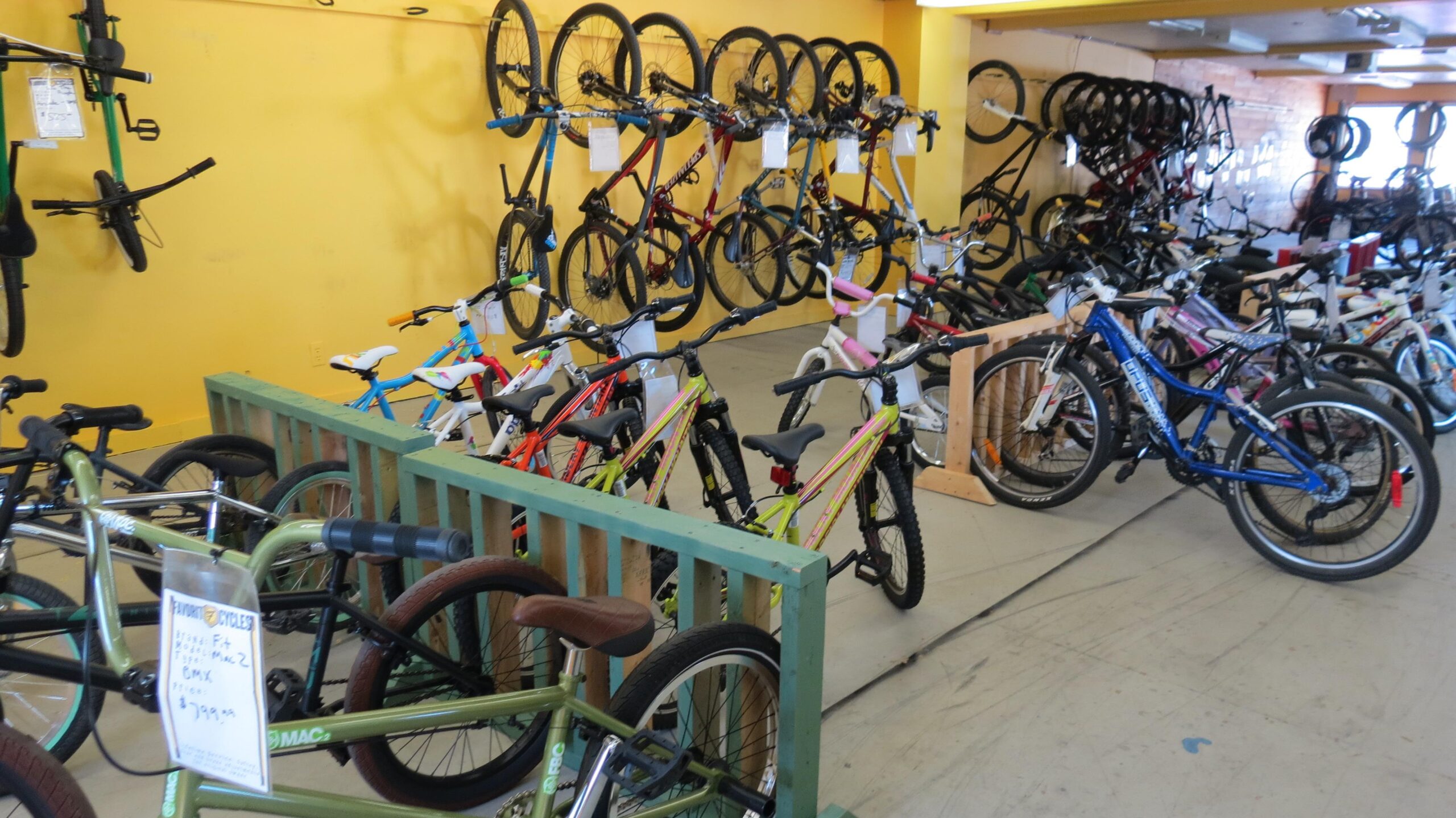 A brightly lit bike shop interior featuring a variety of bicycles. Several bikes are displayed on a wooden rack in the foreground, while additional bikes are mounted on the wall in the background. The shop has yellow walls and a clean floor, with price tags visible on some of the bicycles.