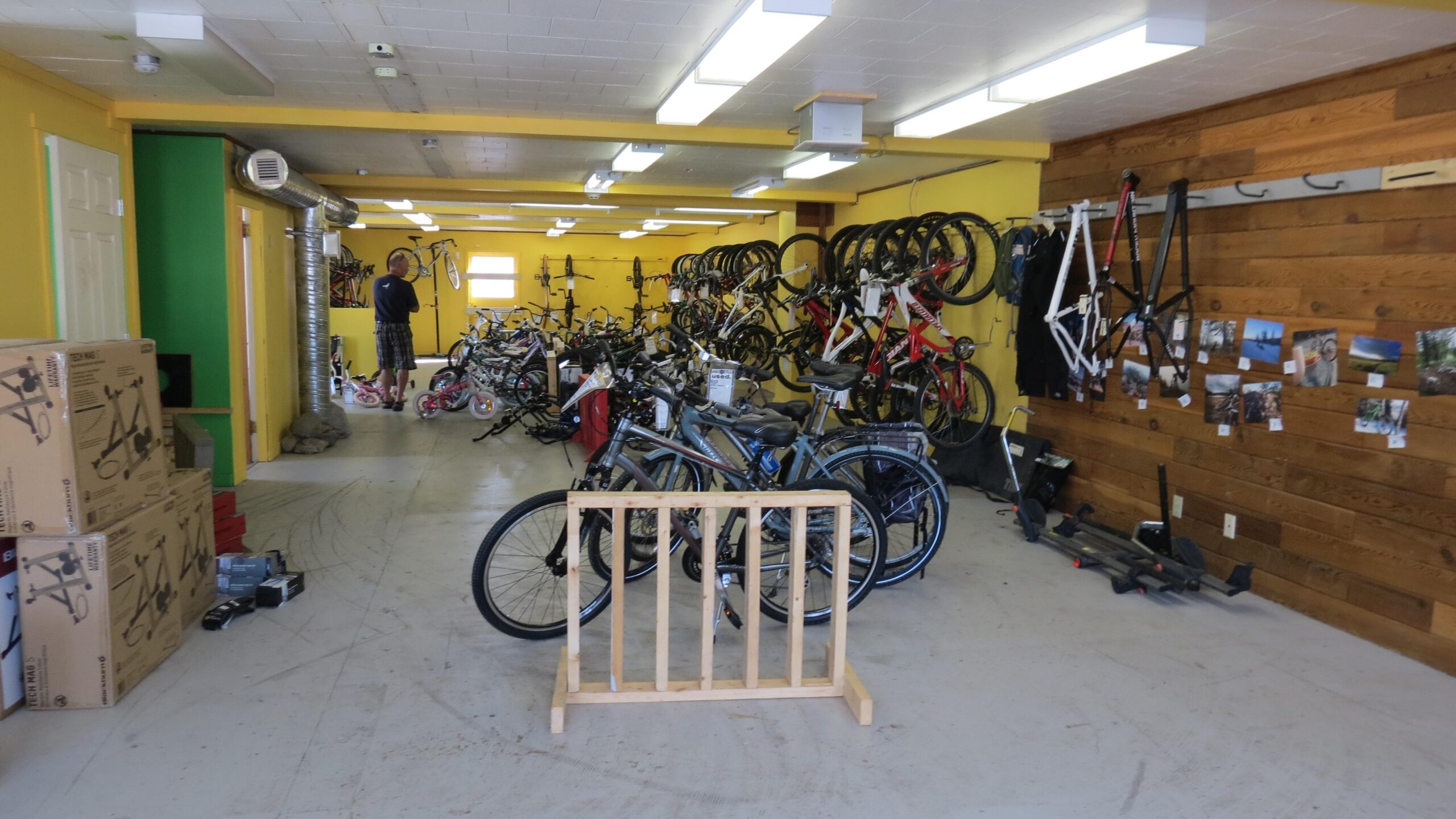 A brightly colored bike shop interior featuring multiple bicycles displayed on the floor and hanging on the walls. The walls are painted yellow with a green accent, and there's wooden paneling on one side. Several cardboard boxes and bike parts are scattered on the ground. In the background, a person is browsing the bicycles.