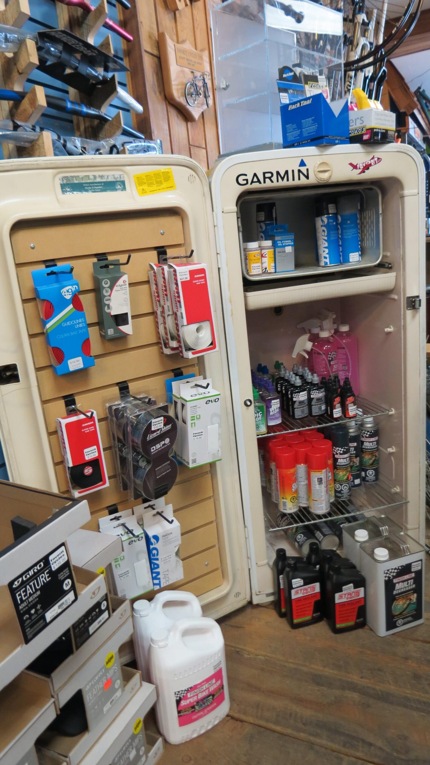 A display in a retail setting featuring a vintage-style refrigerator labeled "Garmin." The fridge is filled with various bicycle maintenance products, including sprays and oils. To the left, a pegboard showcases additional items like cables and accessories, all arranged for easy access. The flooring is wooden, contributing to the overall rustic atmosphere.