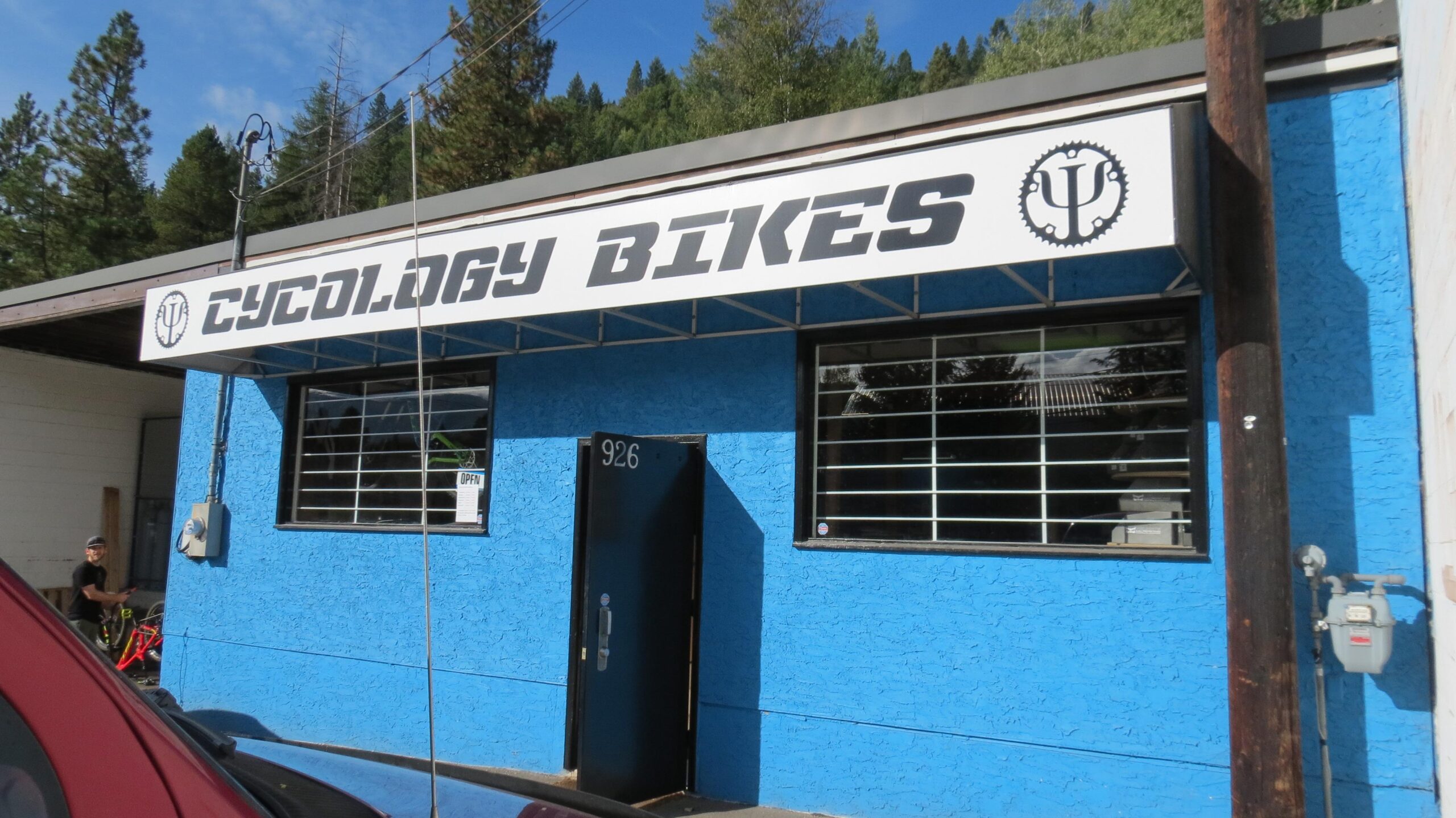A bright blue building with a sign that reads "Cycology Bikes," featuring a large bike gear graphic and an open door. The front has multiple windows. A person is visible near a bicycle outside the shop. Trees are in the background under a clear sky.