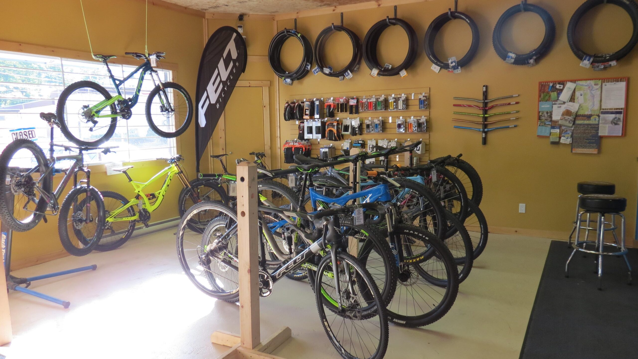 Interior of a bike shop featuring a variety of mountain bikes displayed on the floor and hanging from the ceiling. The walls are lined with bike tires and accessories, including tools and maintenance items. A "Felt" banner is visible, and there are also informational flyers and a bulletin board on one wall.