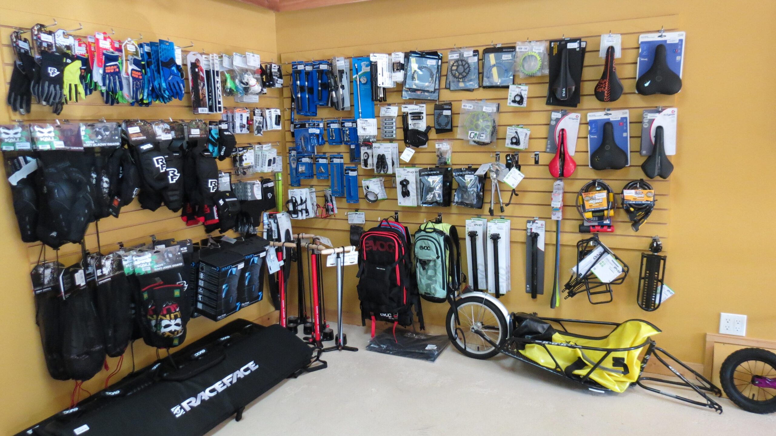 A display wall featuring various cycling accessories and protective gear, including gloves, knee pads, bike seats, pumps, and repair tools. There are also backpacks and a bike trailer on the floor, all set against a yellow background.