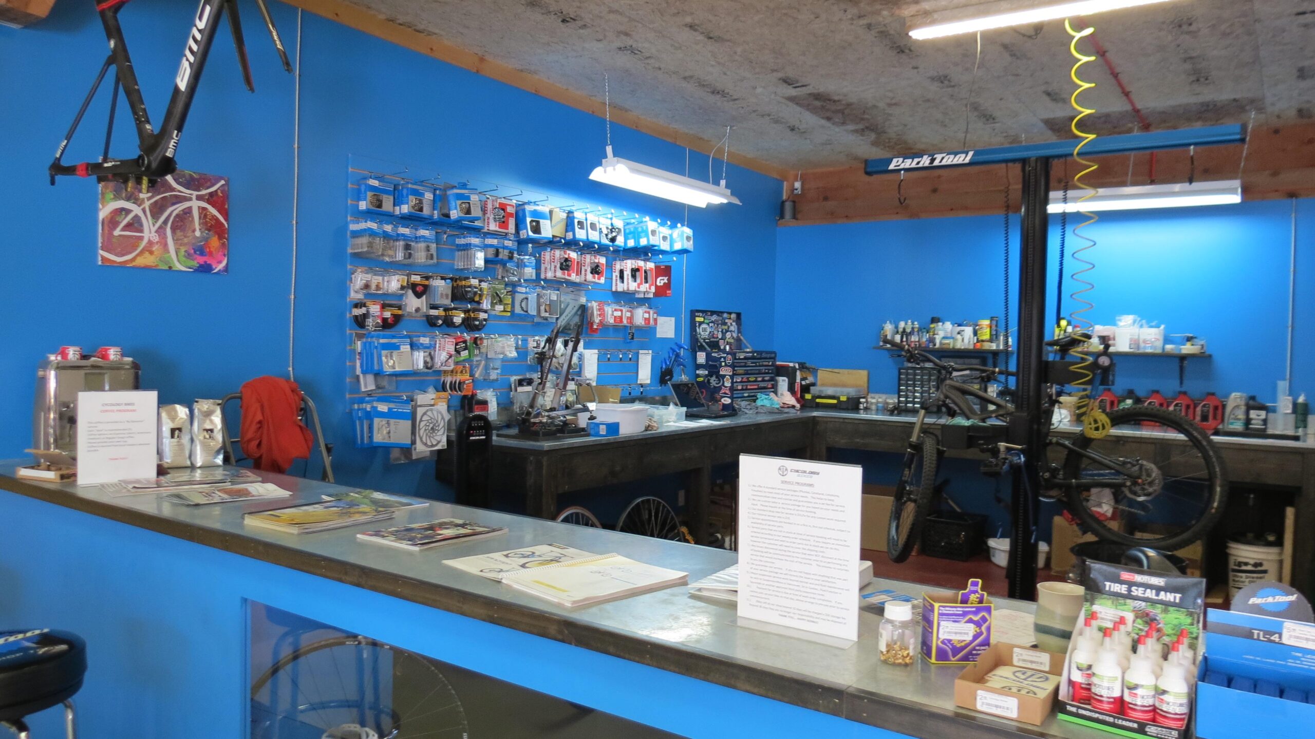 A bright blue interior of a bicycle shop featuring a workbench with tools and bicycle repair equipment. A bike frame is displayed on the wall, and various cycling accessories are organized on shelves. A counter is visible in the foreground with informational materials and a small coffee station.