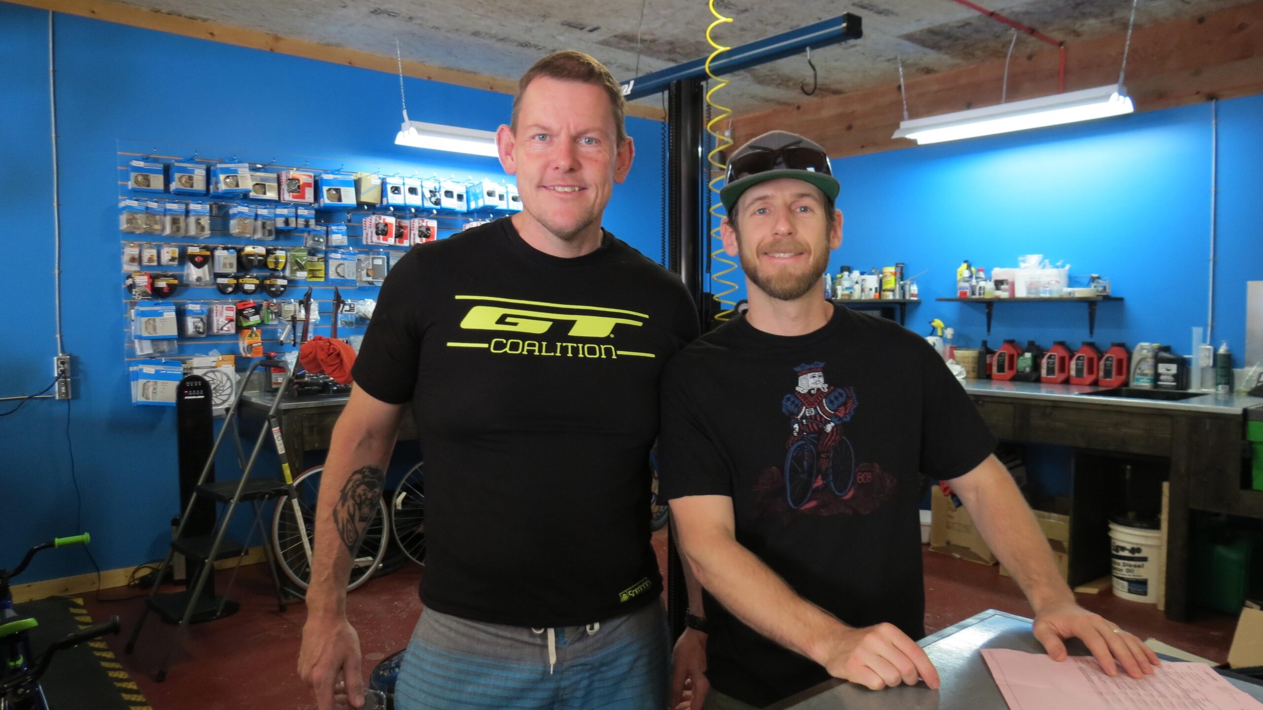 Two men smiling in a bike shop with a blue wall in the background. The man on the left wears a black t-shirt with a yellow logo and has short hair. The man on the right wears a black graphic t-shirt and a green cap. Various bicycle parts and tools are displayed on the wall behind them, and there are supplies and equipment visible on a workbench.