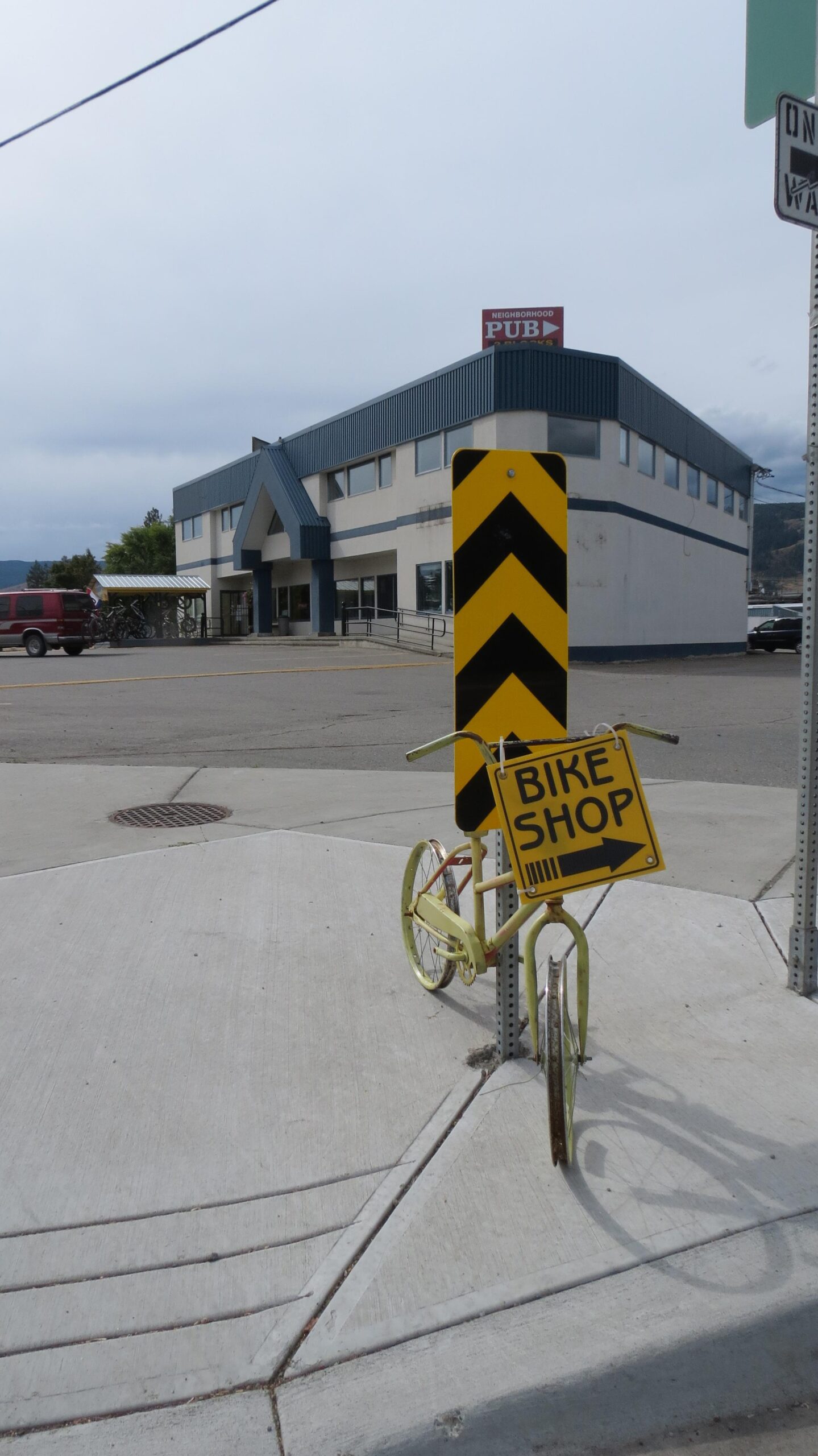 A yellow bicycle with a sign reading "BIKE SHOP" points to the right, situated on the corner of a sidewalk. In the background, a two-story building with a blue roof and large windows is visible, alongside several bicycles parked underneath a shelter. A traffic sign and a nearby road indicate the presence of a bustling neighborhood area.