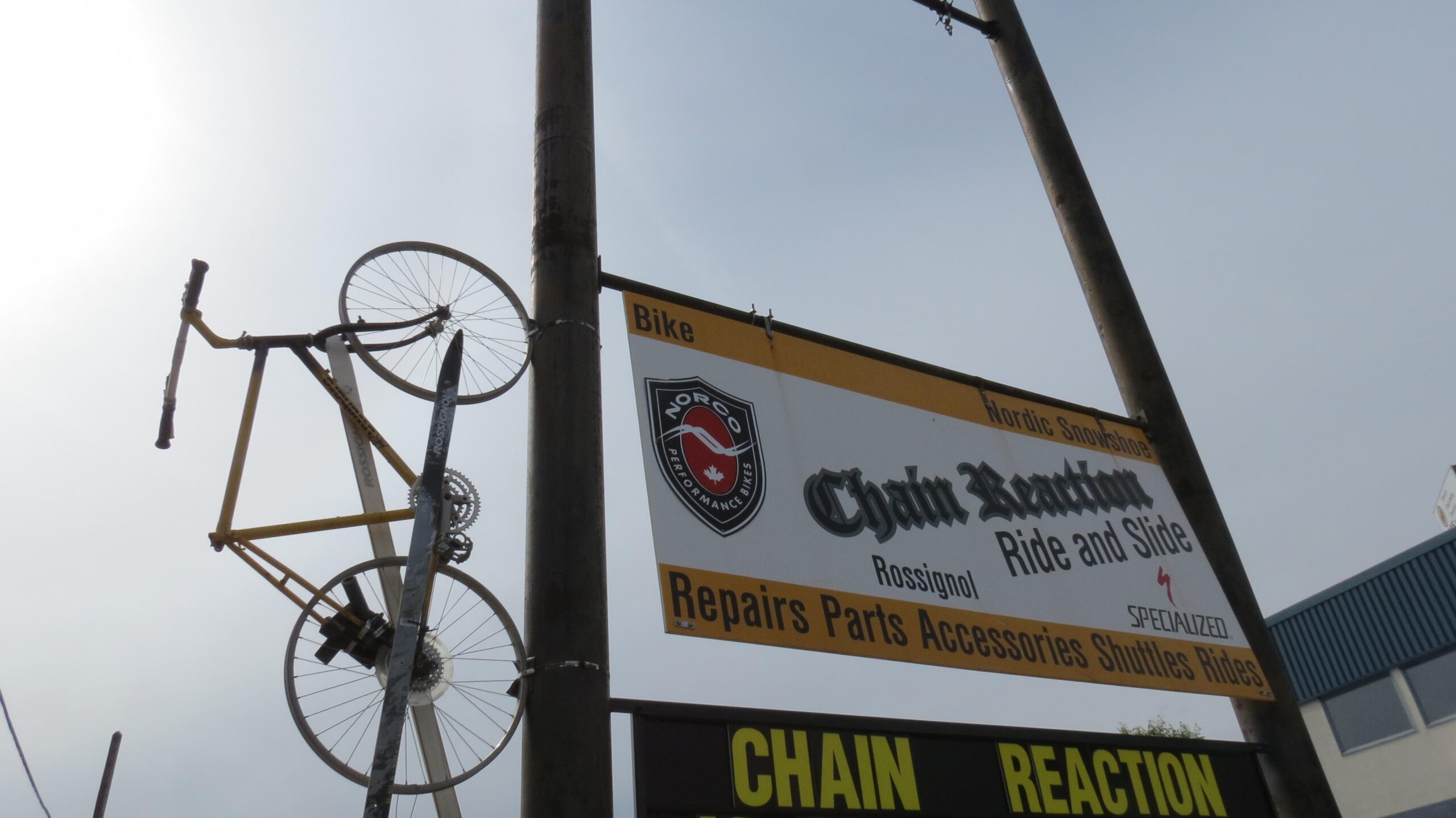 A tall sign mounted on a pole featuring the text "Chain Reaction Ride and Slide," with additional details about bike repairs and accessories. Above the sign, a yellow bicycle frame and one tire are creatively displayed. The background shows a cloudy sky and part of a building.
