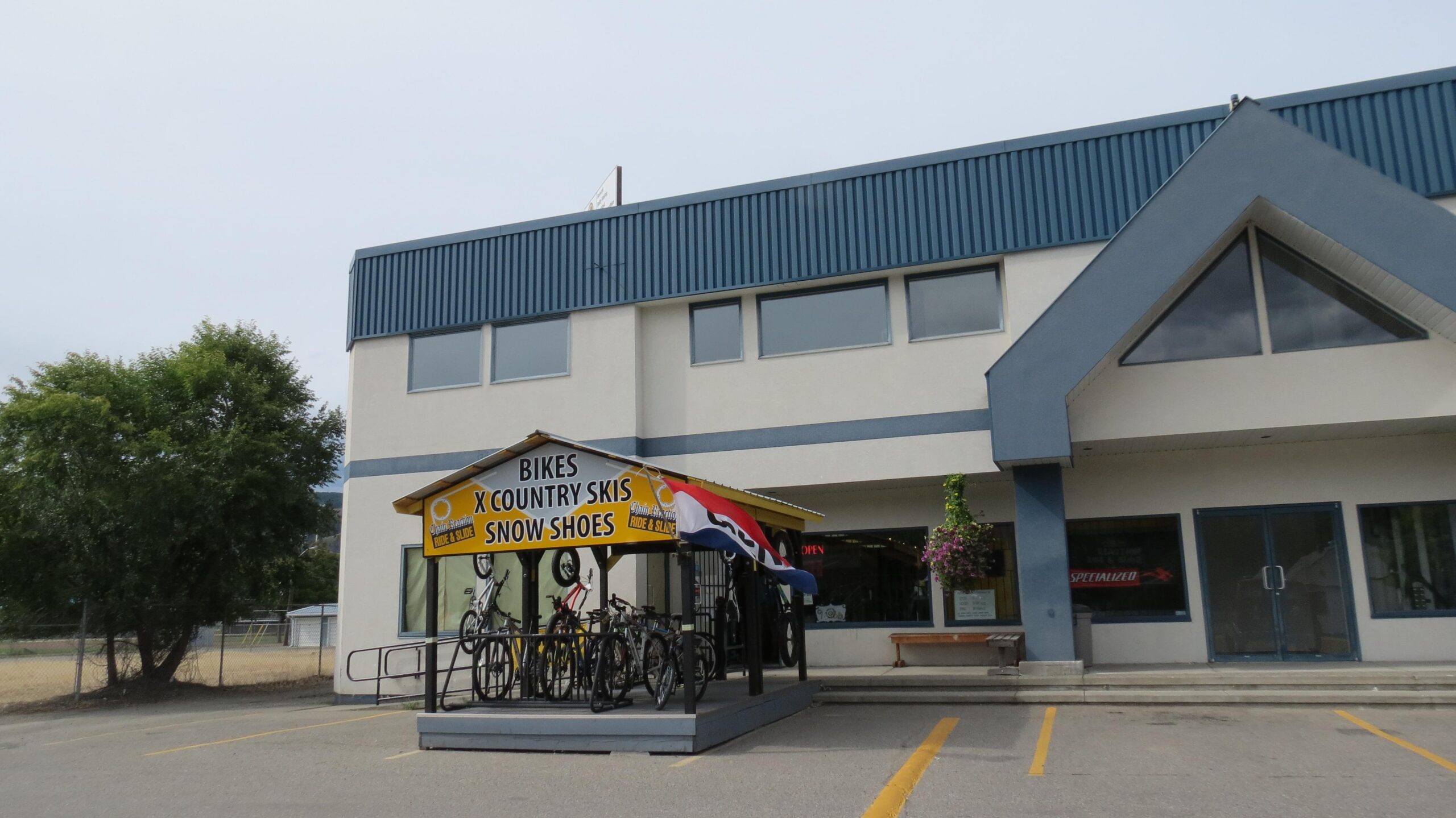 A storefront featuring a colorful awning advertising "Bikes," "X Country Skis," and "Snow Shoes." The entrance includes a display of bicycles, with the facade of a modern building in the background. The sky is overcast, and there are trees nearby.